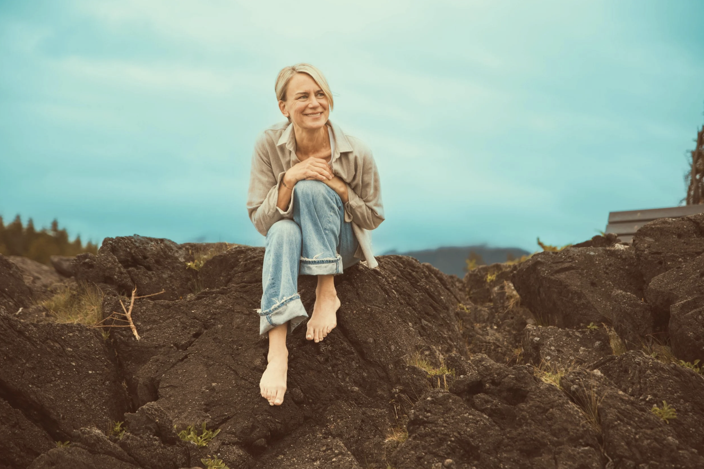 A woman with grey hair and a beige shirt sitting barefoot on rocks outdoors, smiling and gazing upwards with a blue sky and distant mountains in the background.