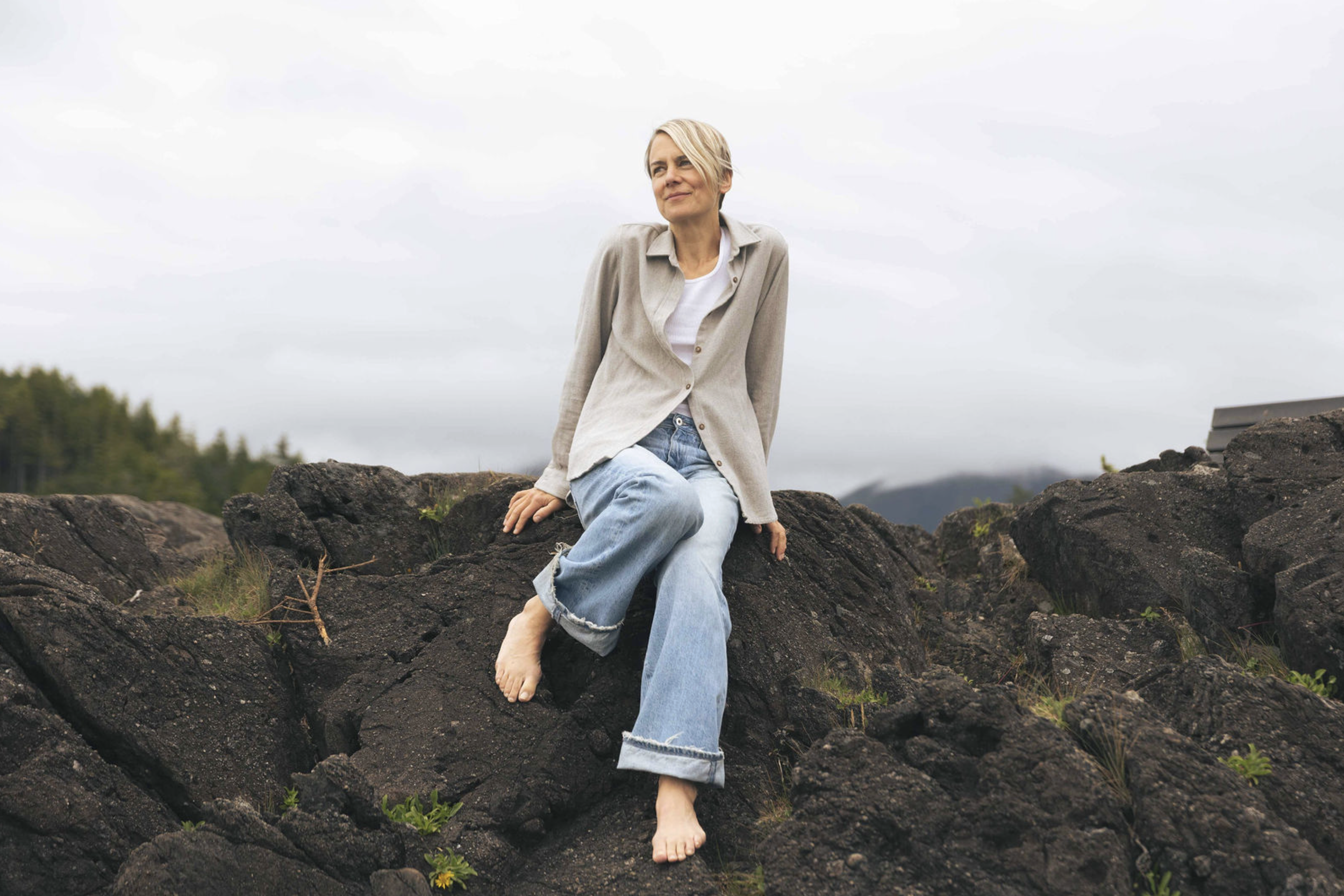 A woman with short blonde hair sitting barefoot on large black rocks outdoors, wearing a beige shirt, white T-shirt, and rolled-up blue jeans. Overcast sky in the background.
