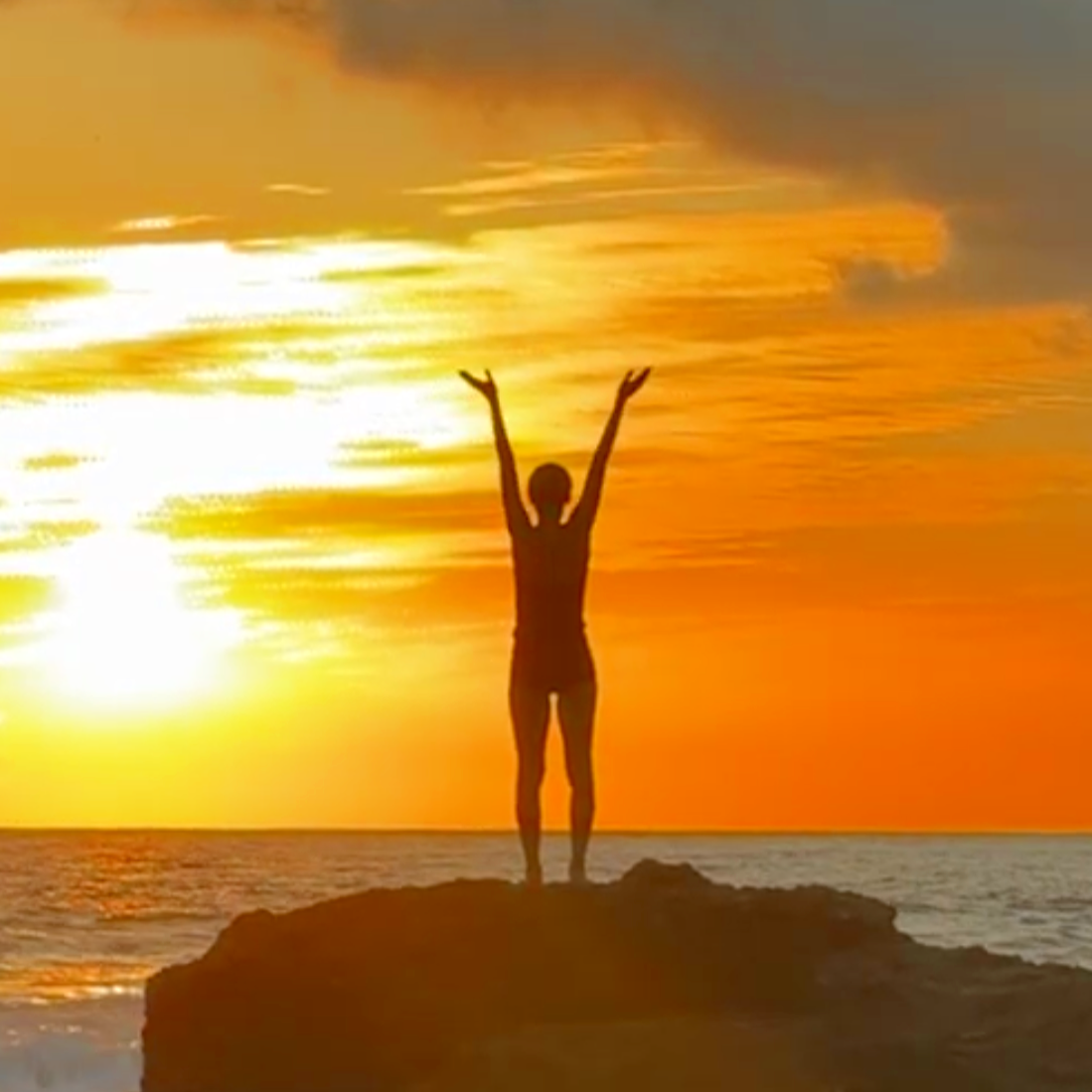 Silhouette of a person standing on a rock with arms raised, facing the sunset over the ocean.