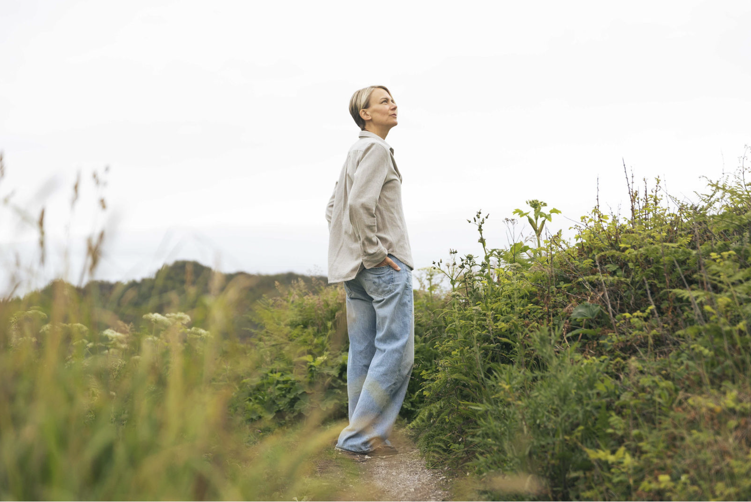 A woman with short blonde hair standing on a dirt path among green bushes and tall grass, looking up at the cloudy sky.