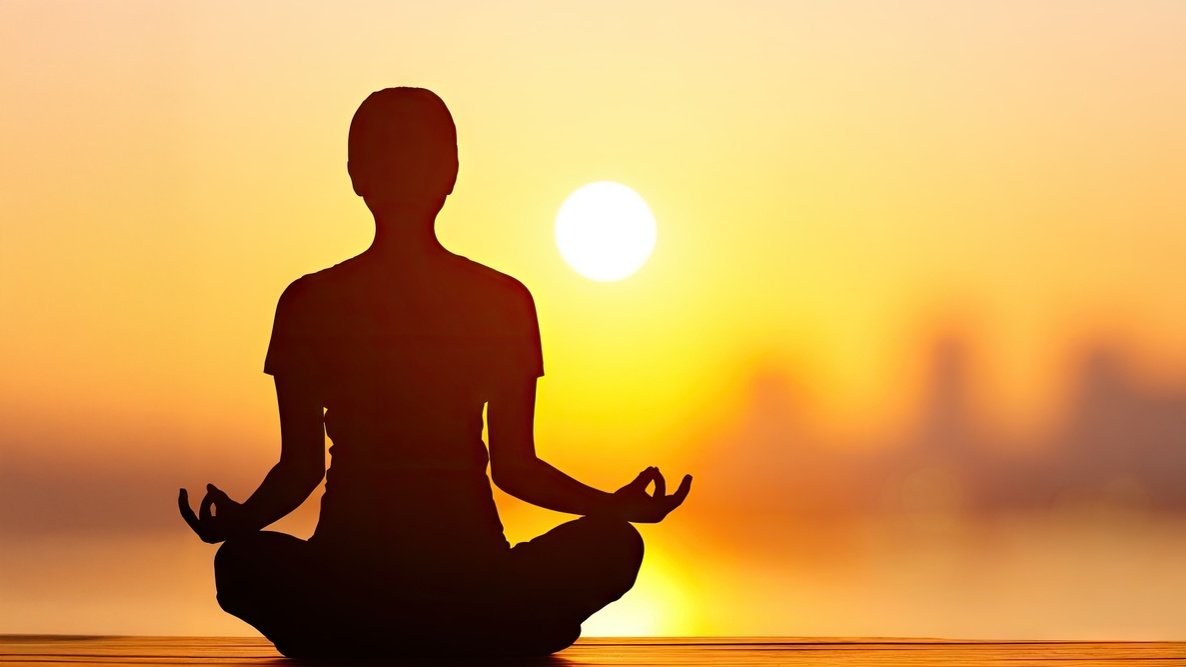 Silhouette of a person sitting in a lotus position practicing meditation at sunset with a calm water background.