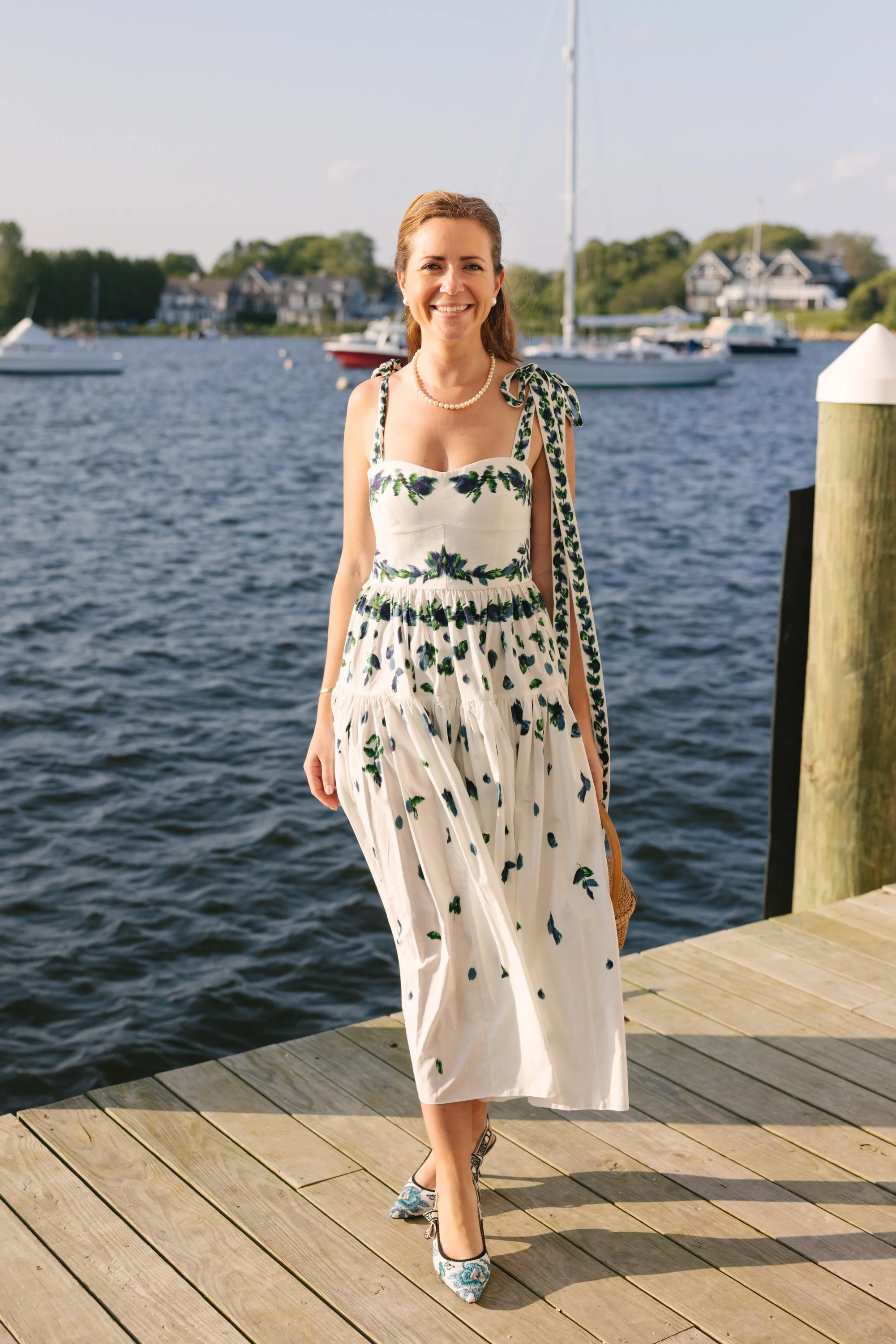 A woman in a floral dress standing on a wooden dock by a body of water with boats and houses in the background, smiling at the camera.