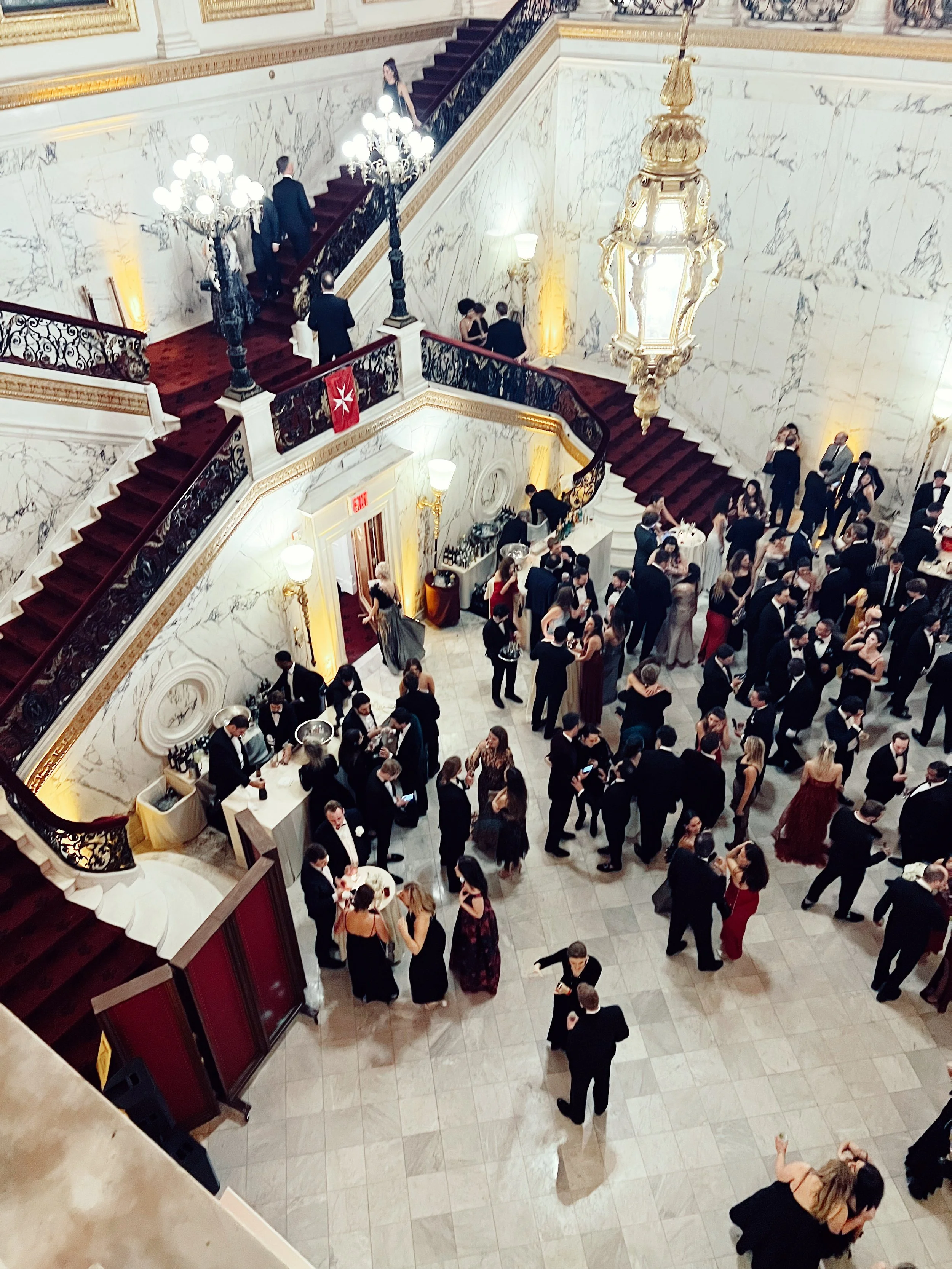 An elegant ballroom filled with formally dressed guests attending a social event or cocktail party. Guests are mingling on the marble floor and ascending the stairs or socializing near the bar. The room features ornate chandeliers, gold accents, and marble walls.