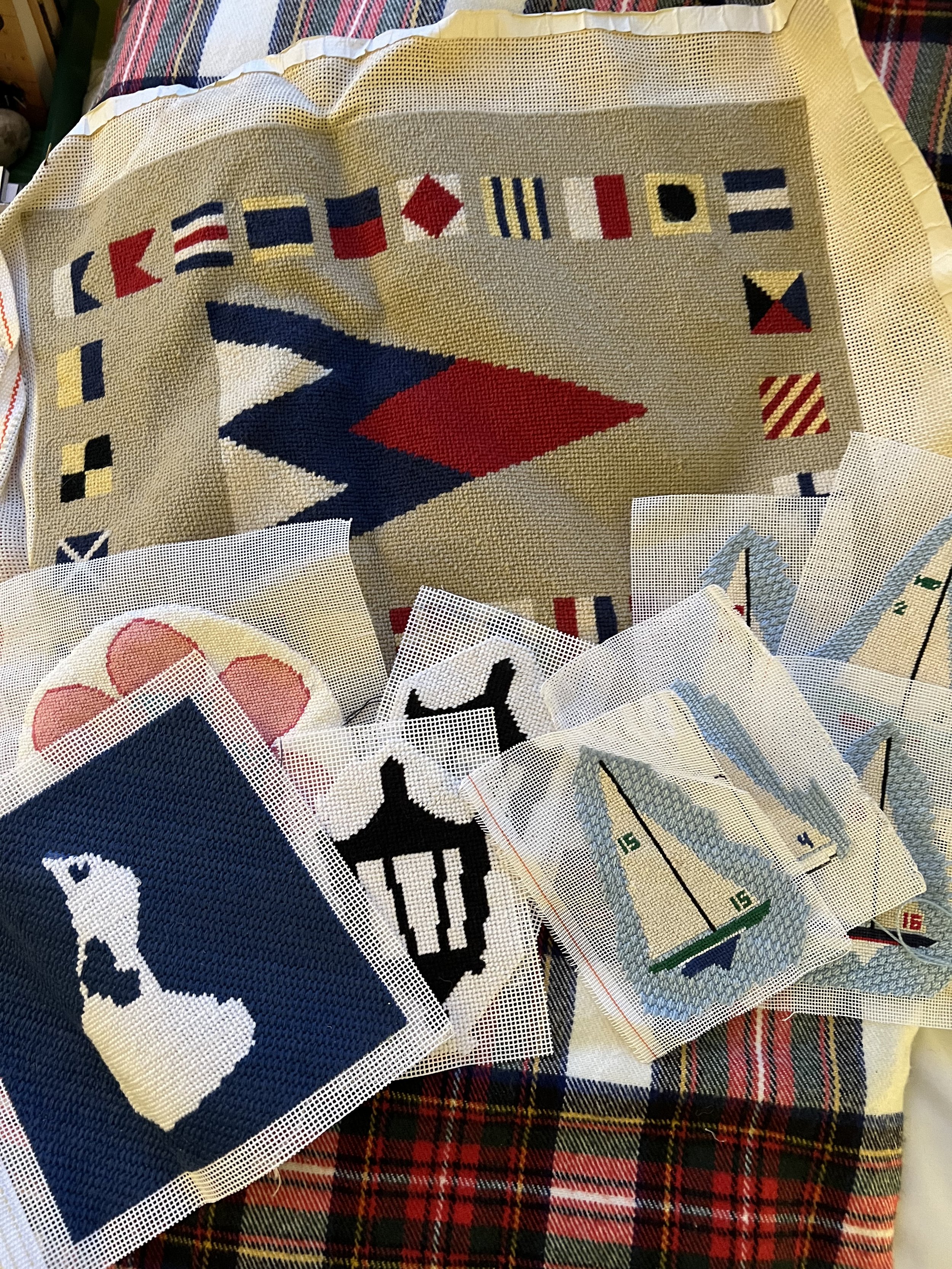 A collection of cross-stitch fabric pieces with various nautical and geometric designs, including flags, a whale, a lighthouse, and a sailboat, spread over a plaid fabric surface.