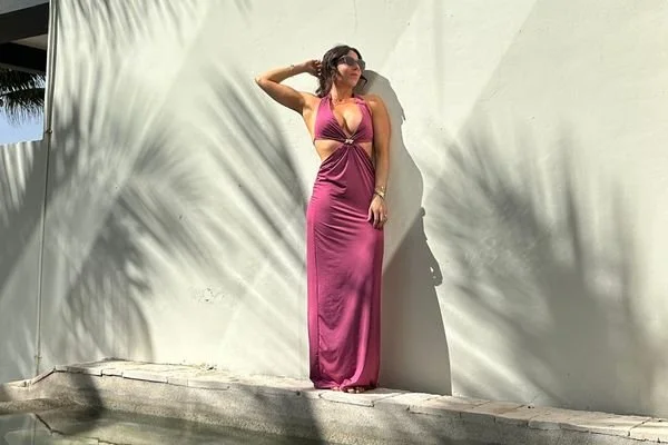 A woman in a long pink dress and sunglasses standing against a light-colored wall with shadows of palm leaves showing off her vacation tan from Pure Luxe Spray Tanning