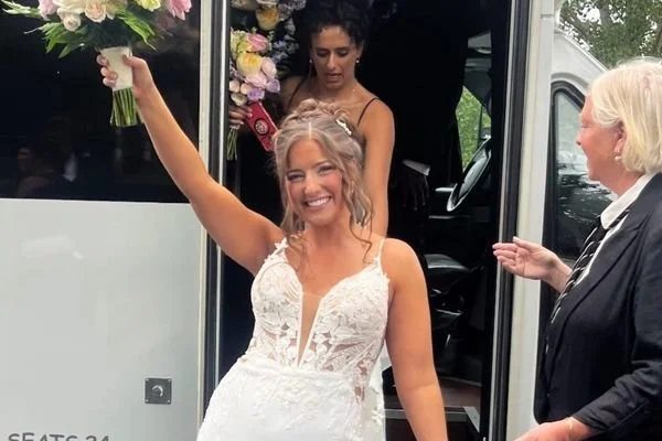 A bride in a white dress holding a bouquet of flowers in one hand, smiling with her arm raised stepping off the bus showing off her flawless bridal tan by Pure Luxe Spray Tanning