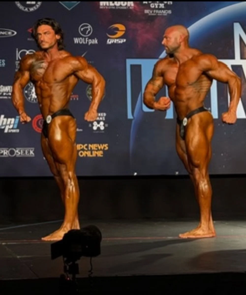Two male bodybuilders are posing on stage during a competition, showcasing their muscular physiques. They are both wearing black posing trunks and are standing in front of a backdrop with various sports and fitness logos.