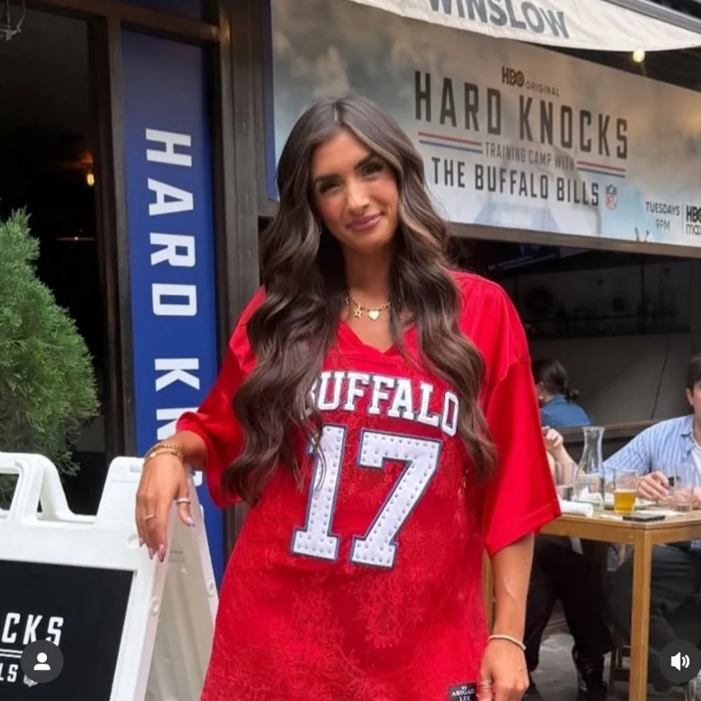 A woman with long wavy hair wearing a red Buffalo Bills football jersey with the number 17 standing outside a restaurant with a spray tan from Pure Luxe