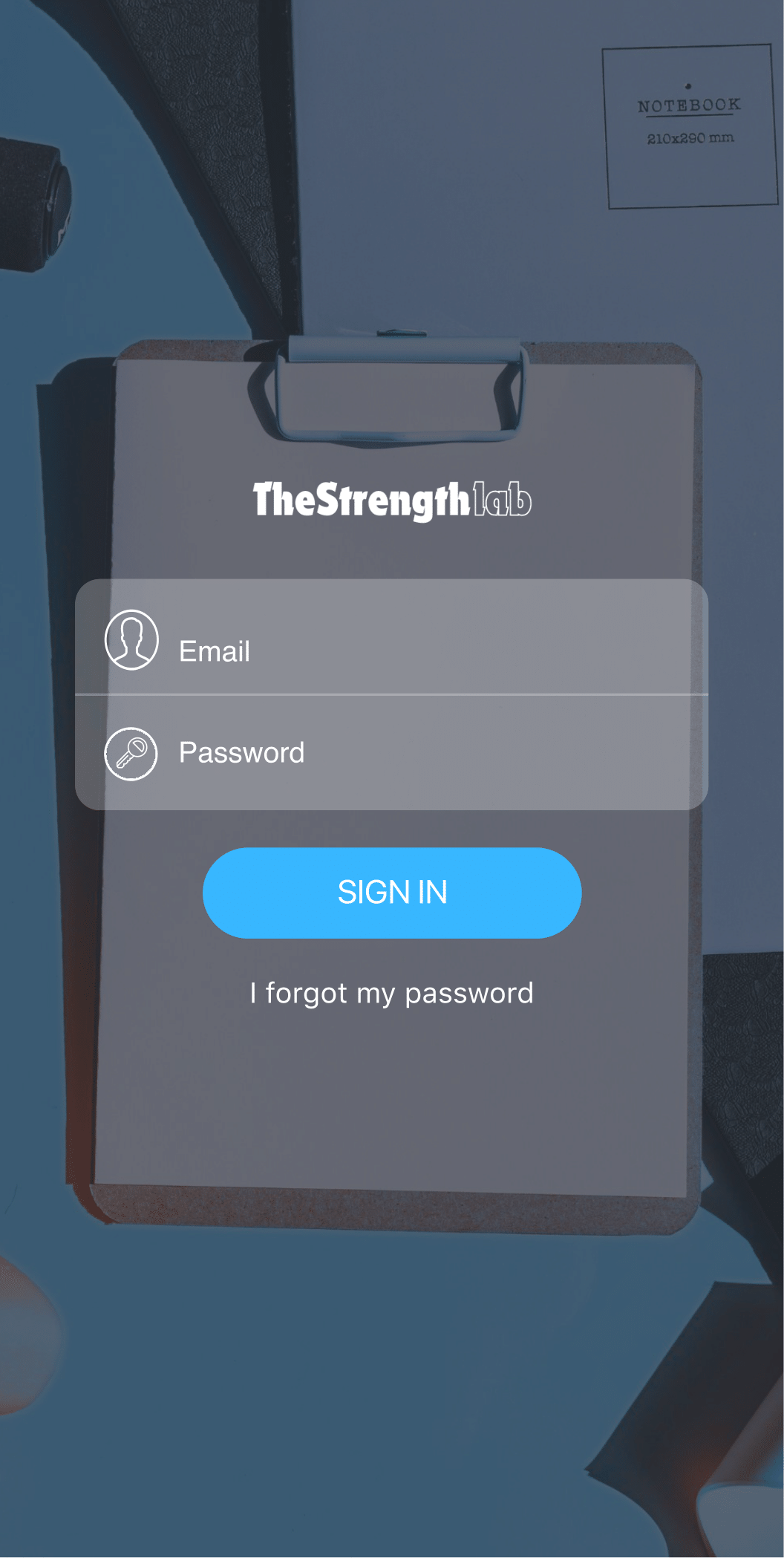 Login screen for The Strength lab app, with fields for email and password, and a blue sign-in button, on a clipboard on a desk with a notebook and other office supplies.