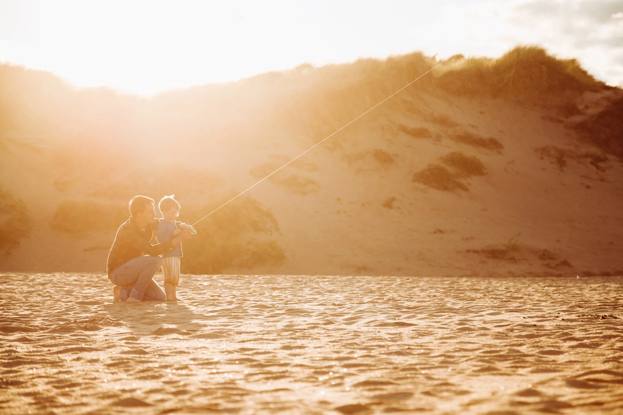 A father and child fishing together on a sandy beach at sunset with dunes in the background.