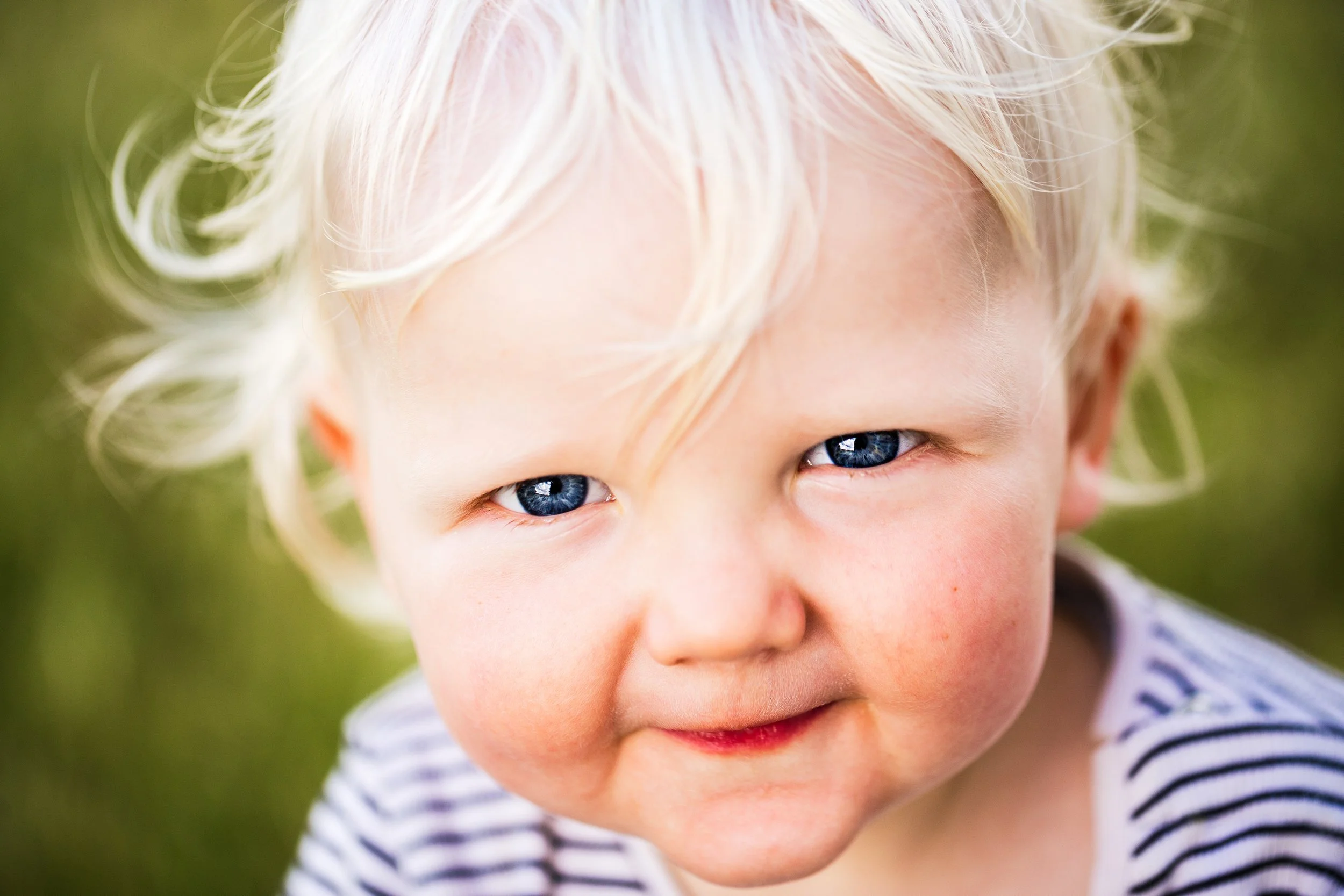 Close-up of a young child with blond curly hair and blue eyes, wearing a striped shirt, smiling outdoors.