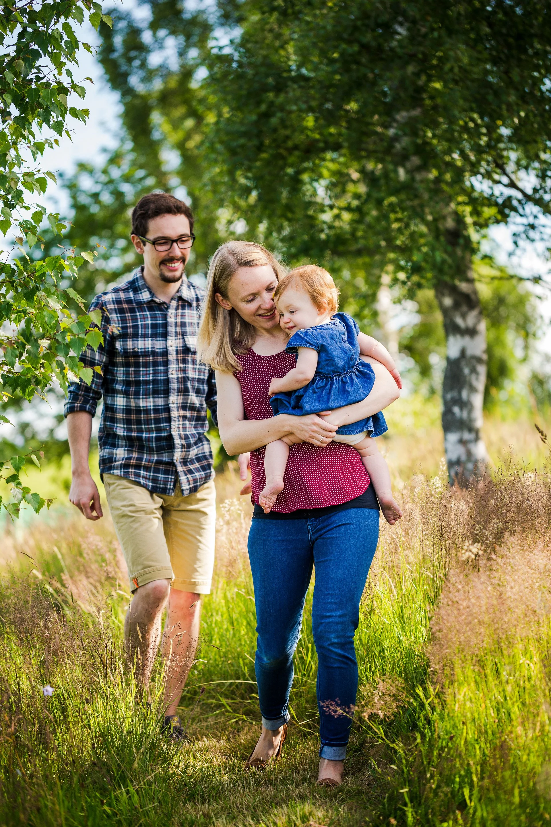 A family of three walking outdoors on a sunny day, with the mother holding a young girl, and the father walking behind them, surrounded by green trees and tall grass.