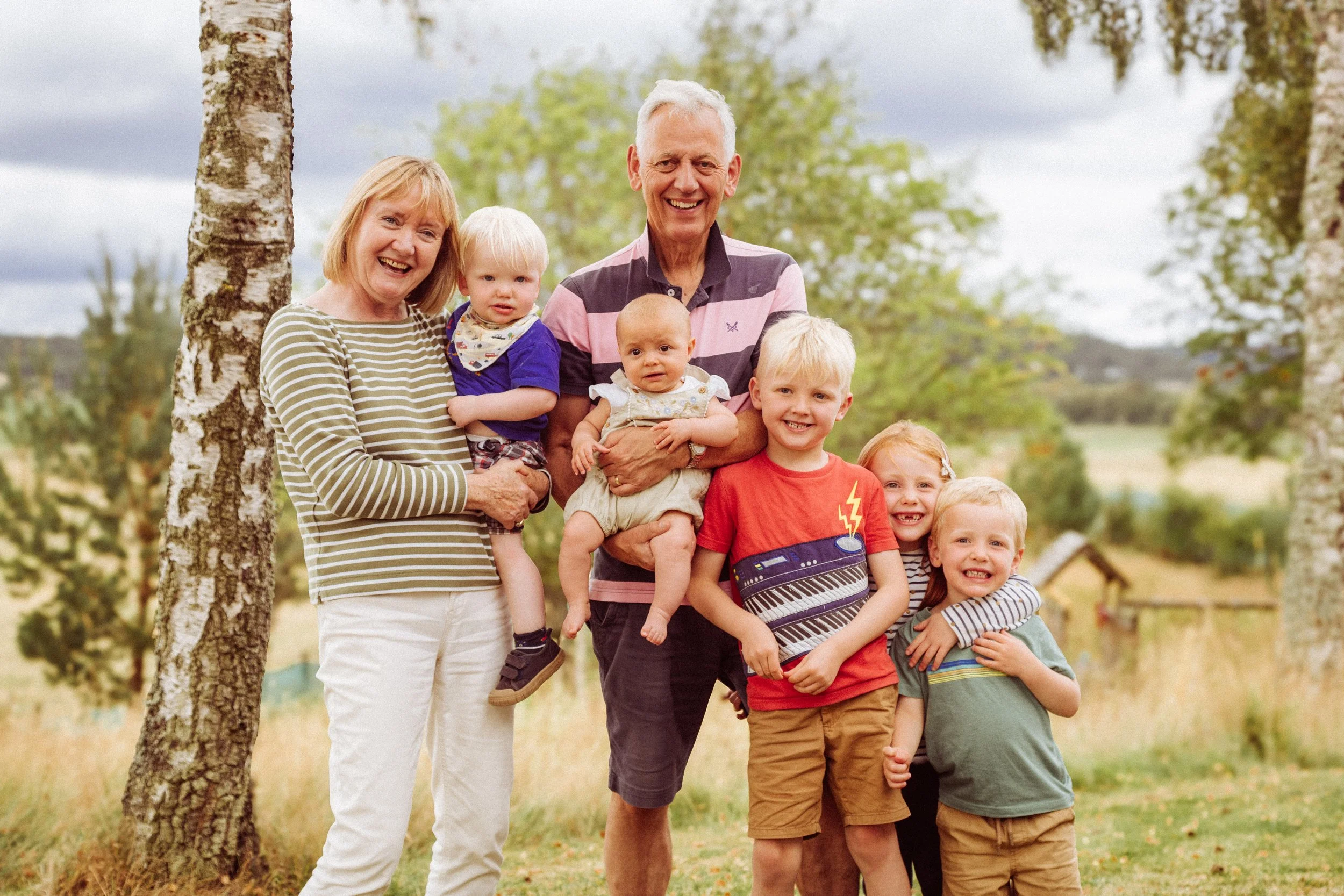 Family photo of two grandparents with five young children outdoors in a park with trees and grass.
