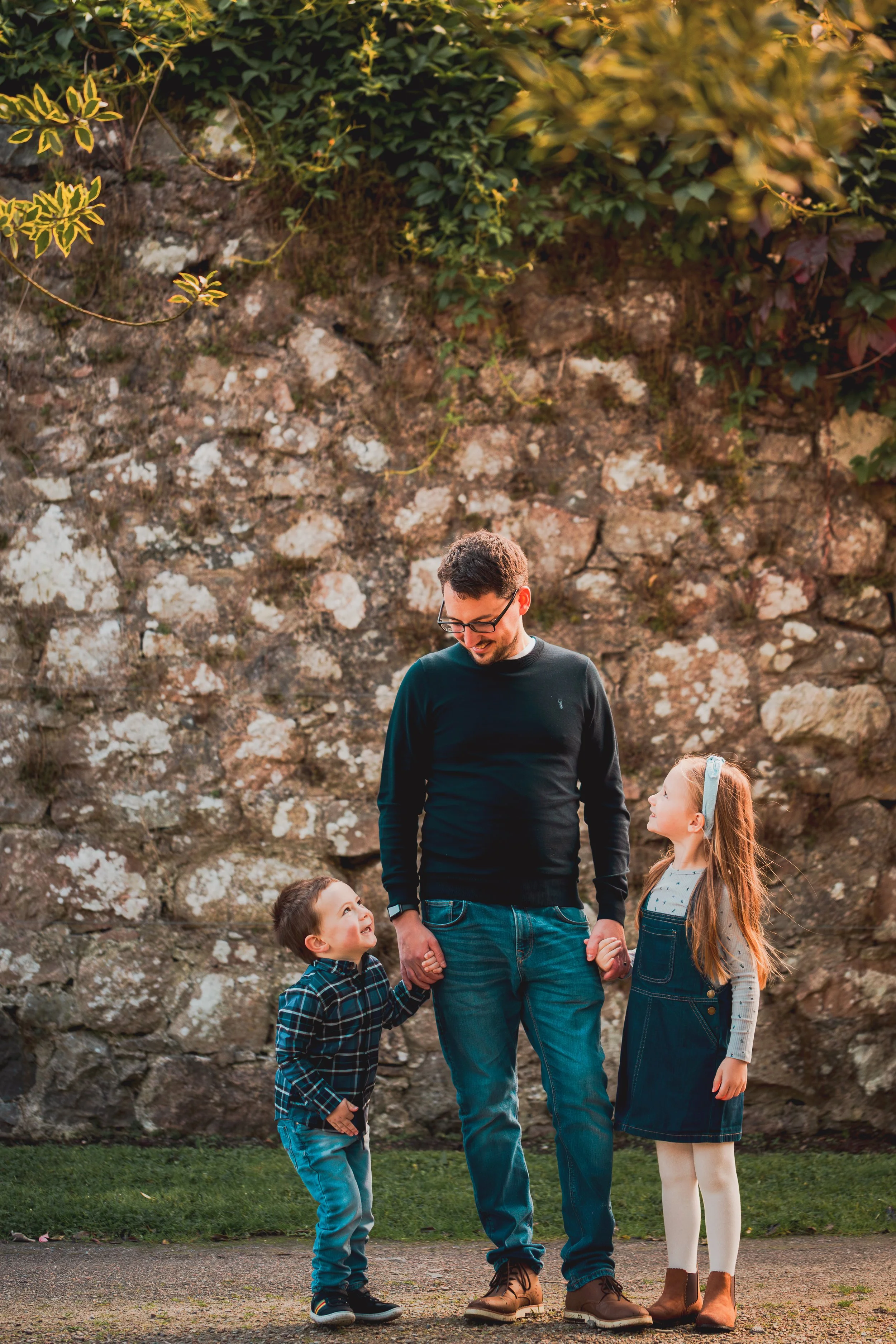 A man holding hands with a young boy and girl, standing outdoors in front of a stone wall with greenery, smiling and looking down at the children.