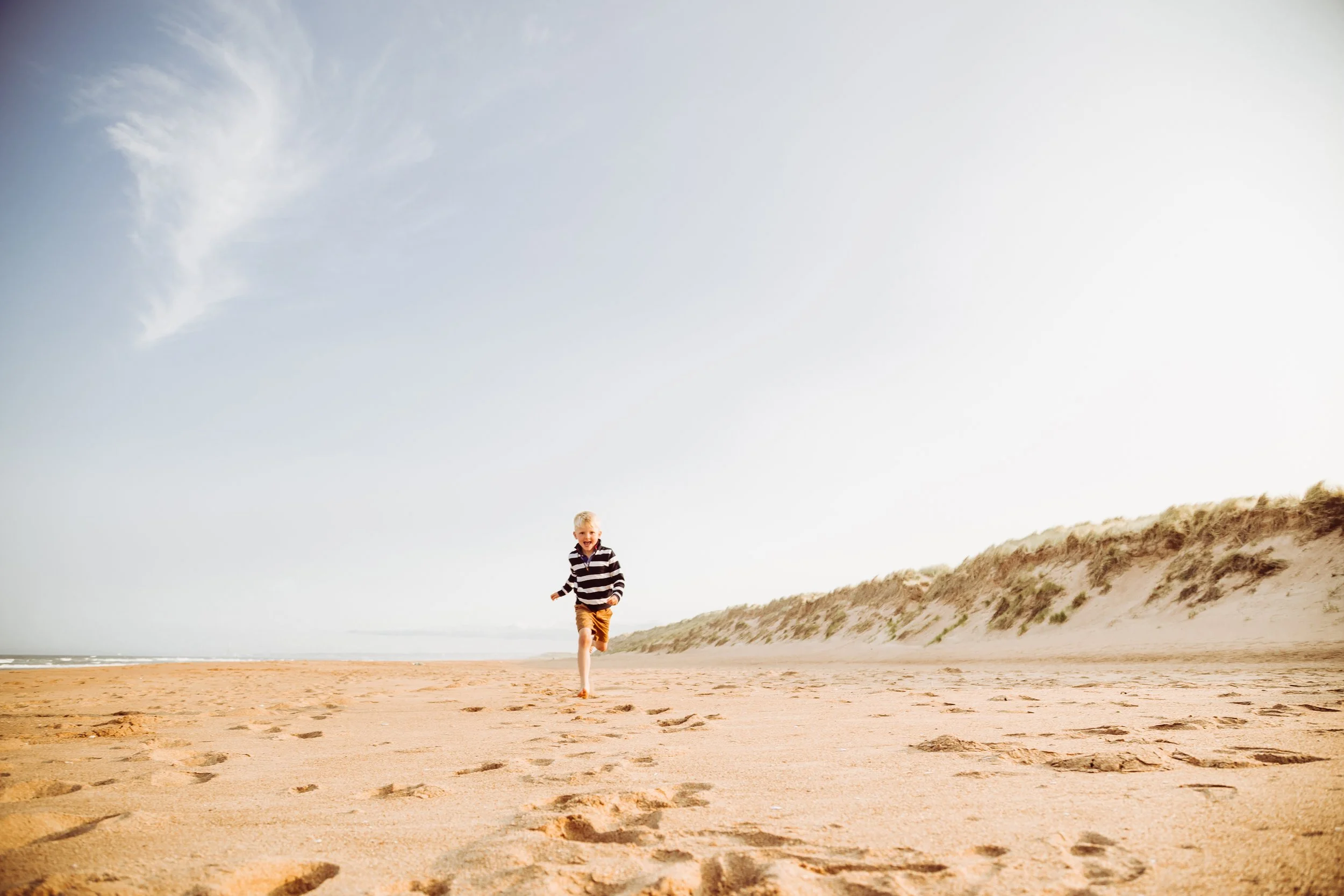 A young boy running on a sandy beach, with sand dunes in the background and a bright sky overhead.
