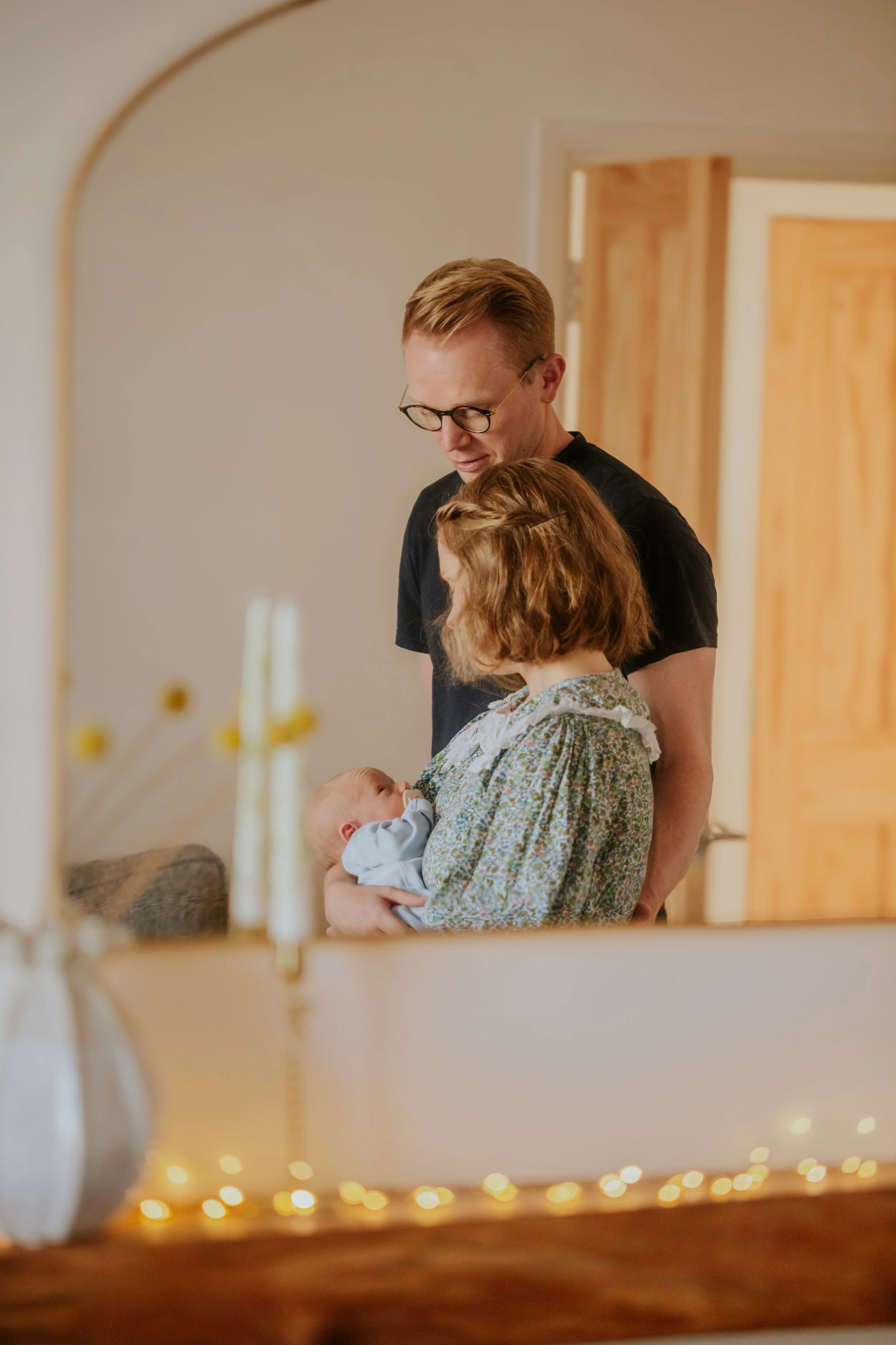 A family of three, including a woman holding a baby and a man standing close, looking at the baby, inside a home with warm lighting and wooden accents.