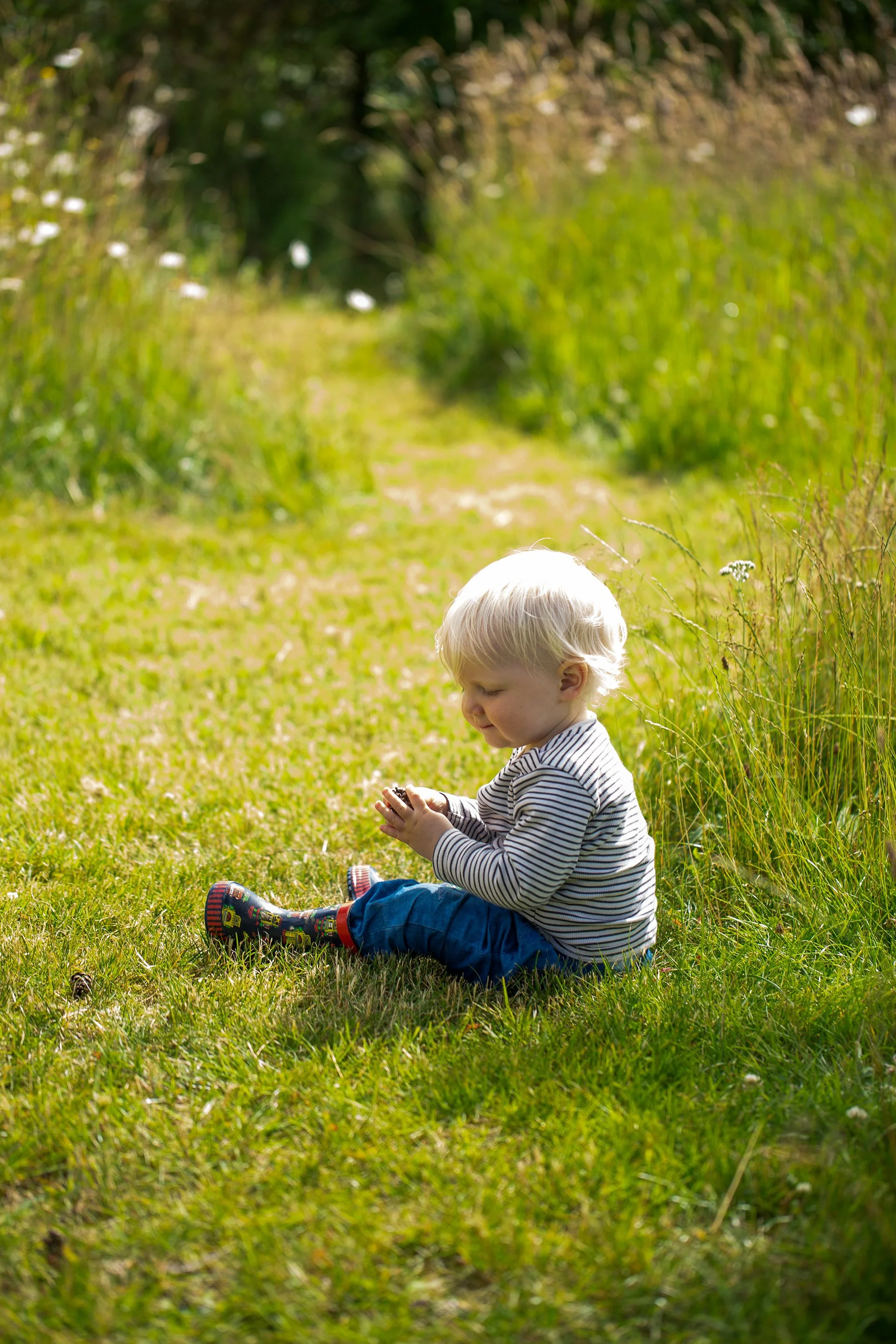 A young child sitting on grass in a garden, playing with a small object, surrounded by green plants and sunlight.