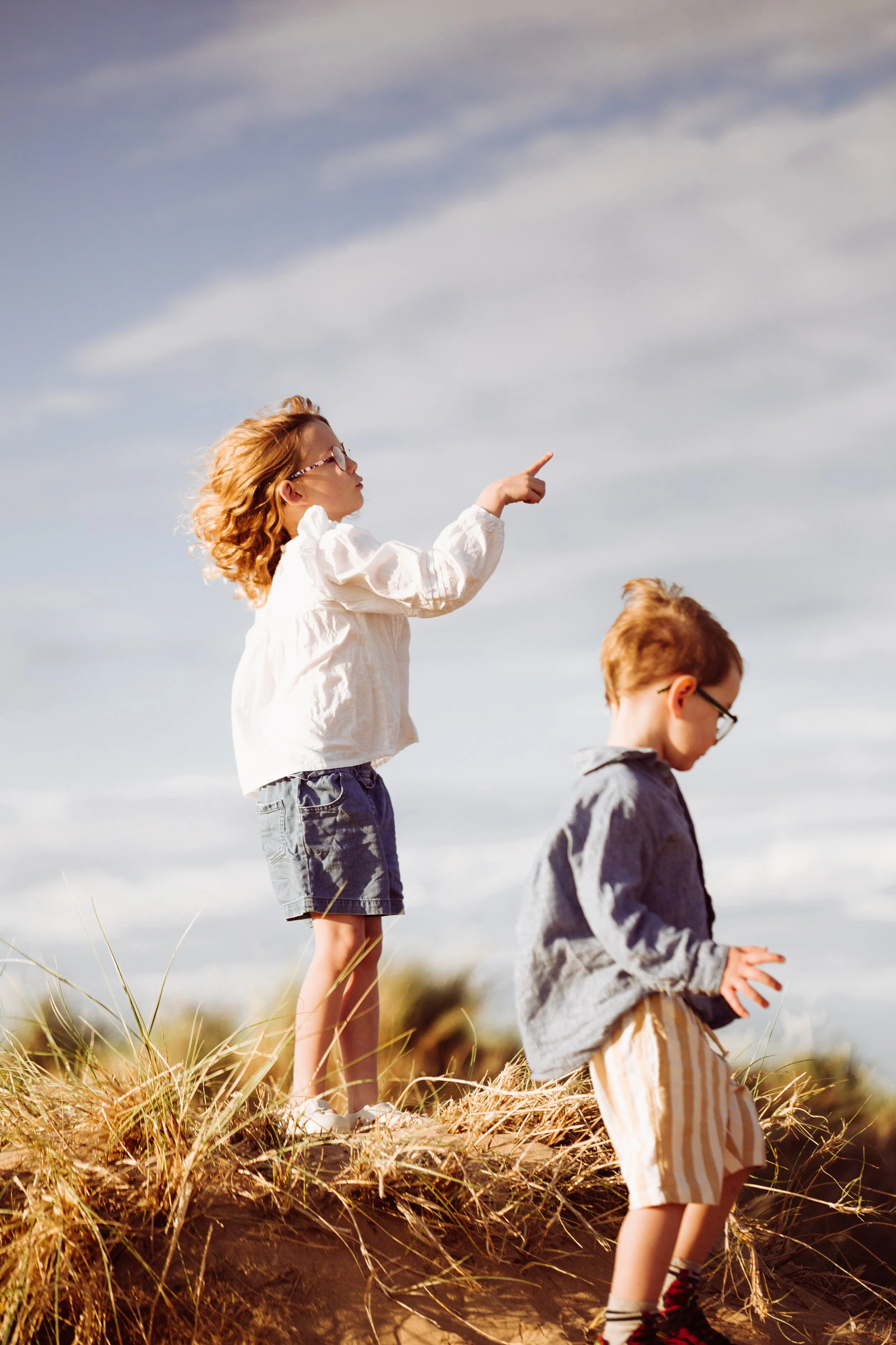 Two children, a girl and a boy, standing outdoors on a grassy hilltop, with the girl pointing into the distance and the boy looking down, under partly cloudy skies.