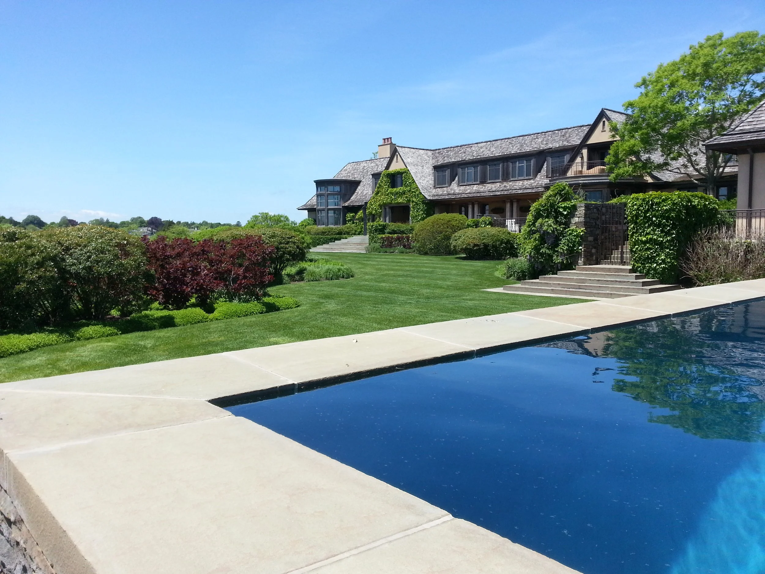 Landscape next to an infinity pool with estate home in background. Home is located South of Highway in Water Mill, NY.