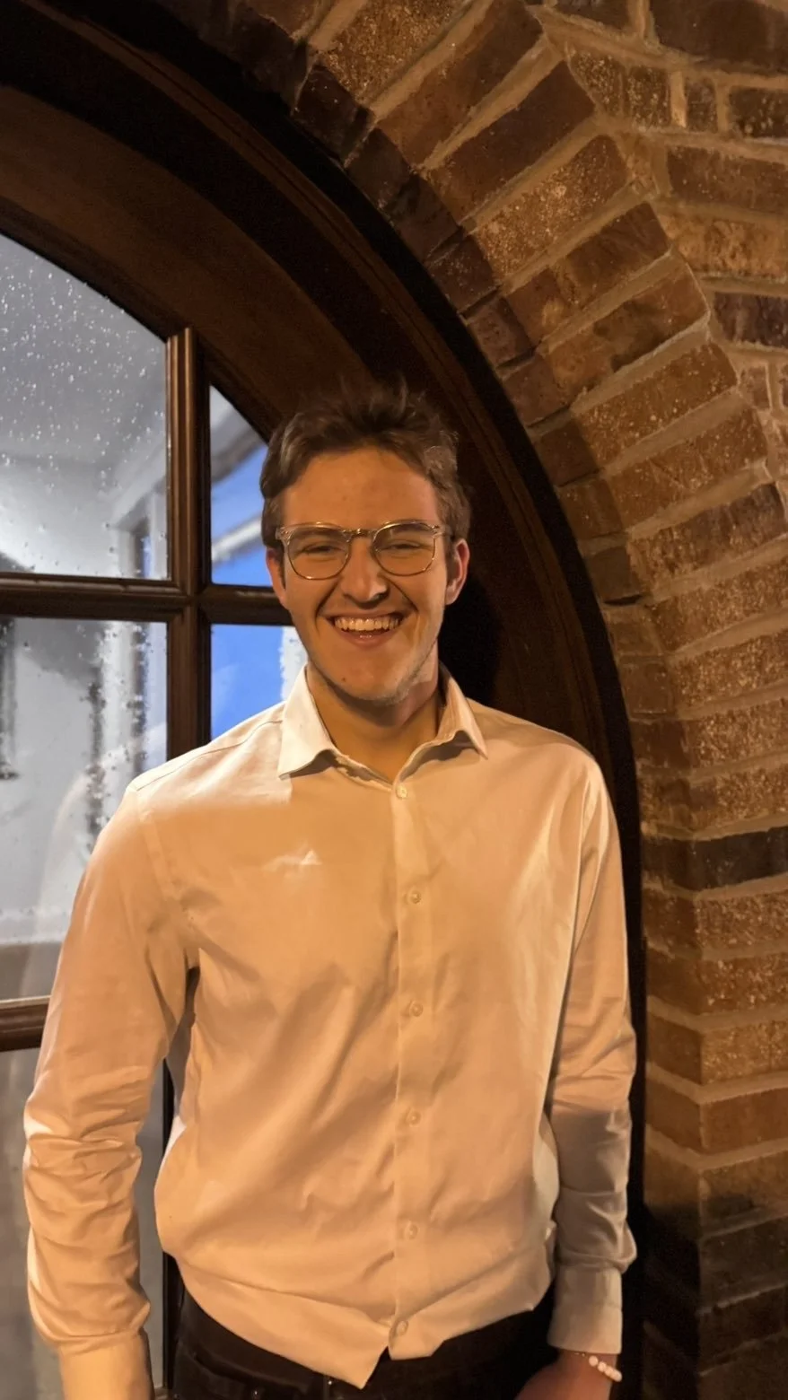 A young man smiling, wearing glasses and a beige button-up shirt, standing in front of a brick archway near a window with rain outside.