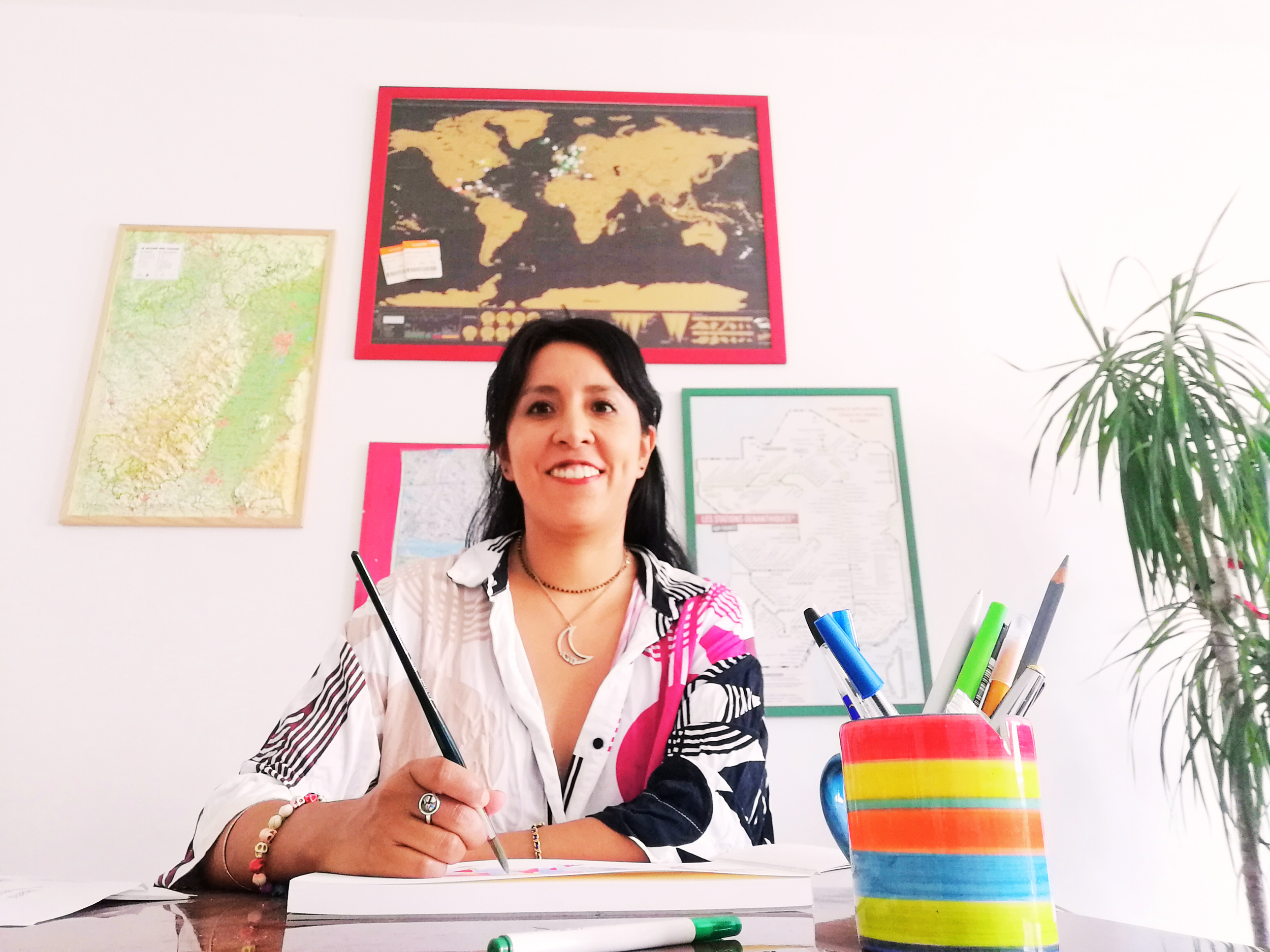 Une femme assise à un bureau, tenant un stylo, avec des cartes et des posters du monde sur le mur derrière elle, et une plante à sa droite.