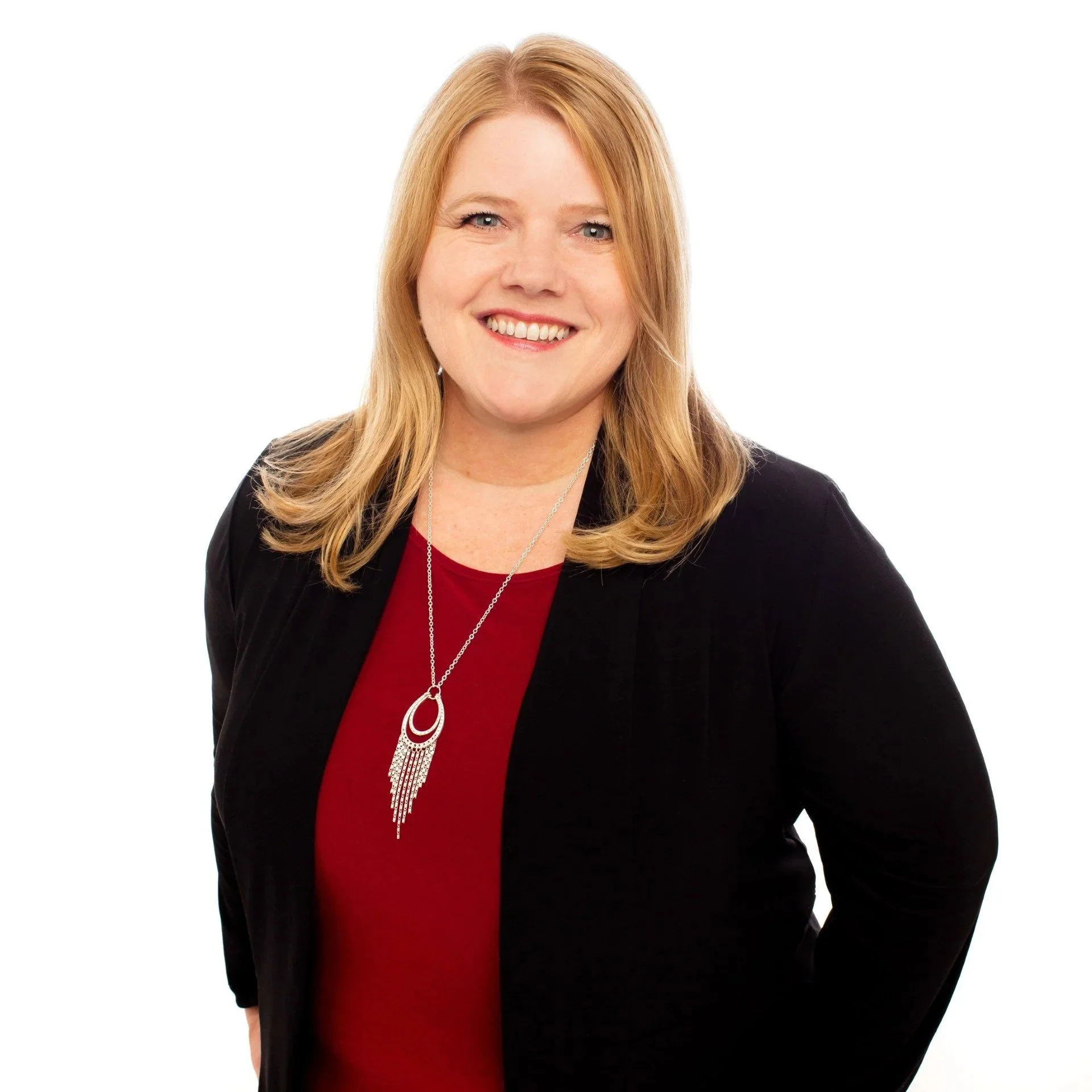 Headshot of a smiling woman with blonde hair wearing a black blazer, red top, and a silver necklace with a pendant, against a white background.