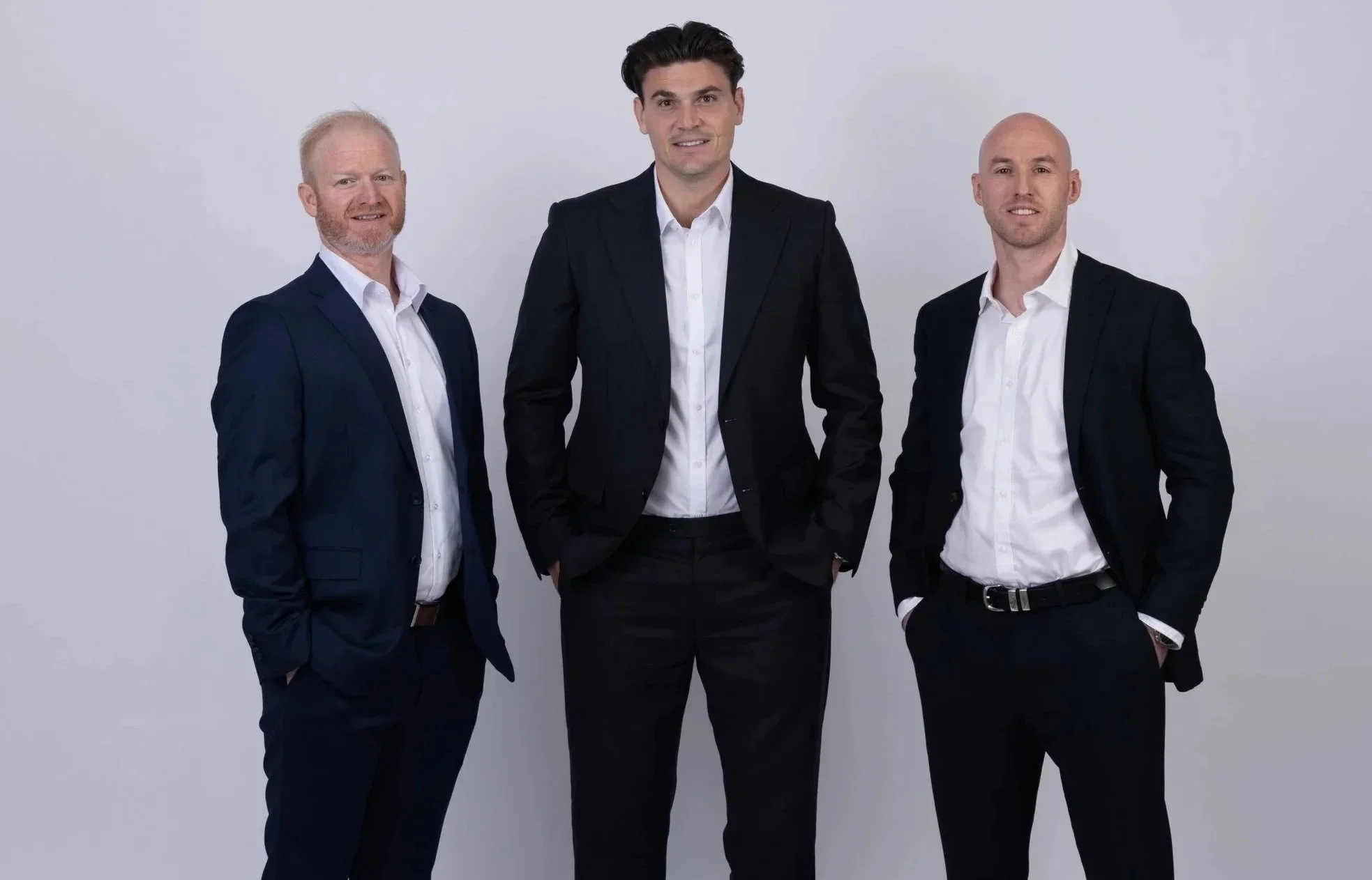 Three men standing together against a plain white background, dressed in dark suits and white shirts, with the man in the center having dark hair and the two men on either side being bald or with light hair.