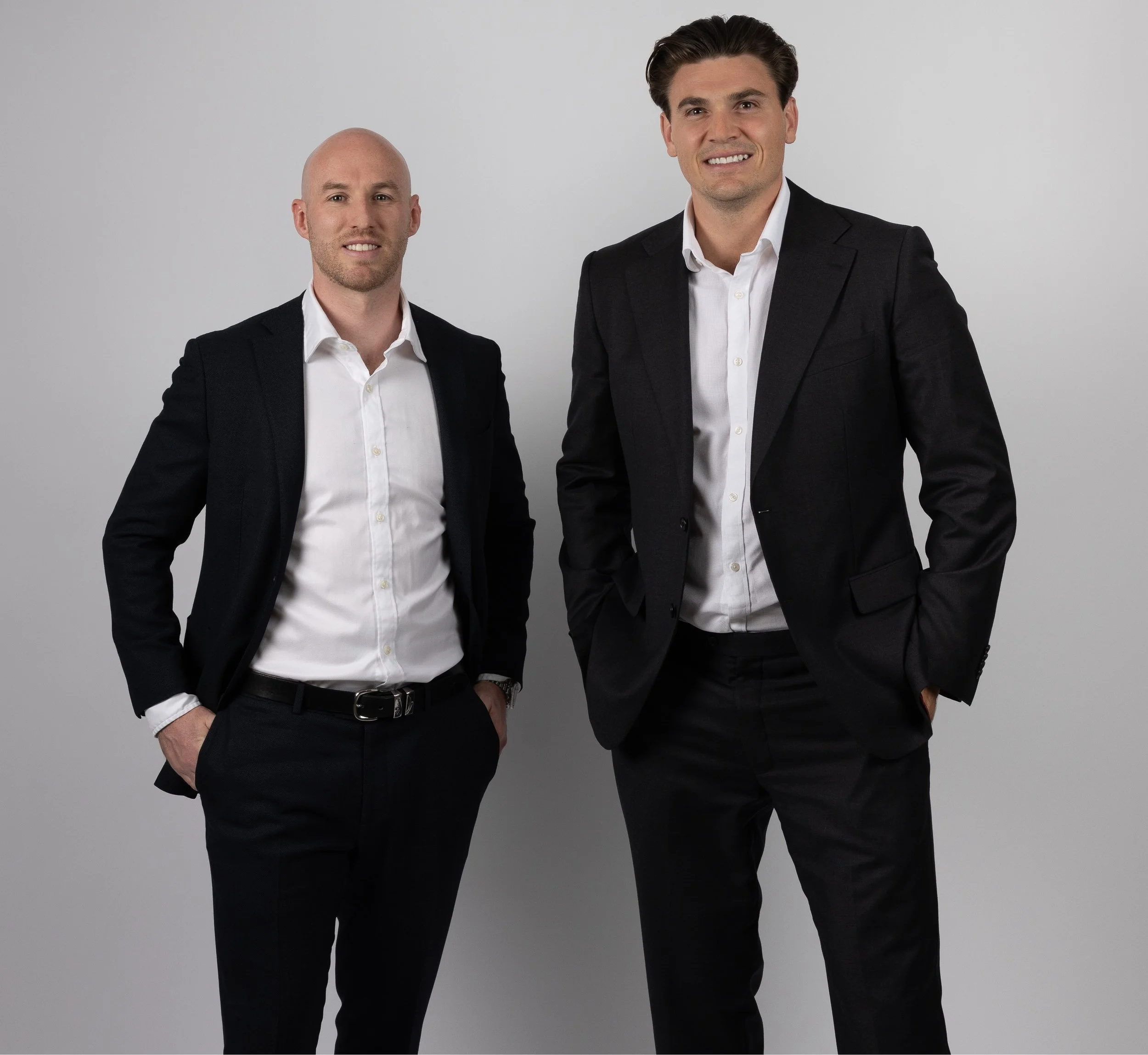 Two men in business suits standing against a plain gray background. The man on the left is bald with a beard, and the man on the right has dark hair and is taller. Both are smiling, hands in pockets, wearing white shirts and dark jackets.