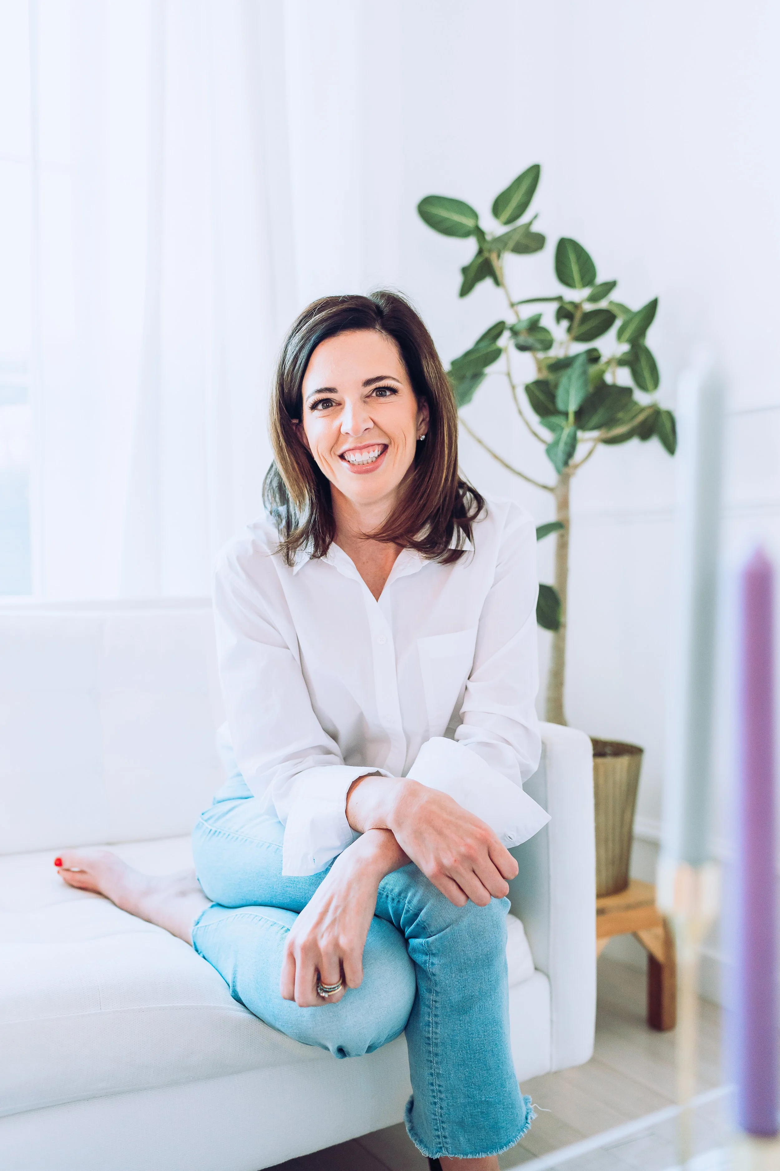 Professional therapist sitting comfortably on a white couch in a light-filled office with greenery, conveying calm, trust, and a welcoming therapy environment.