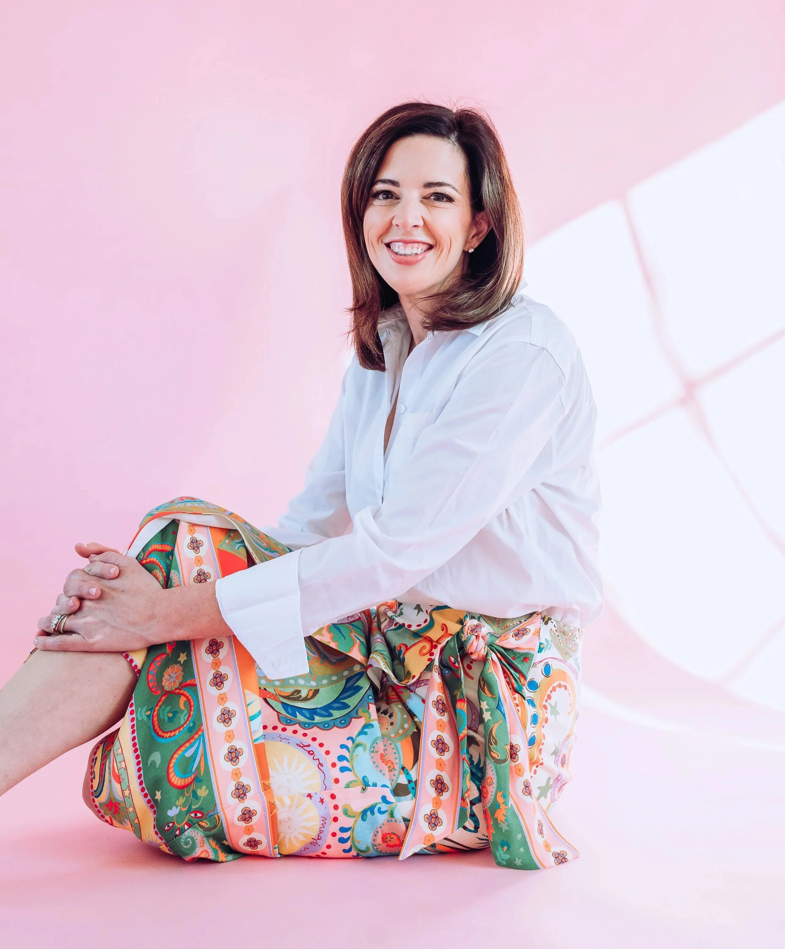 Compassionate therapist seated and smiling in a soft pink studio setting, reflecting warmth, ease, and emotional safety.