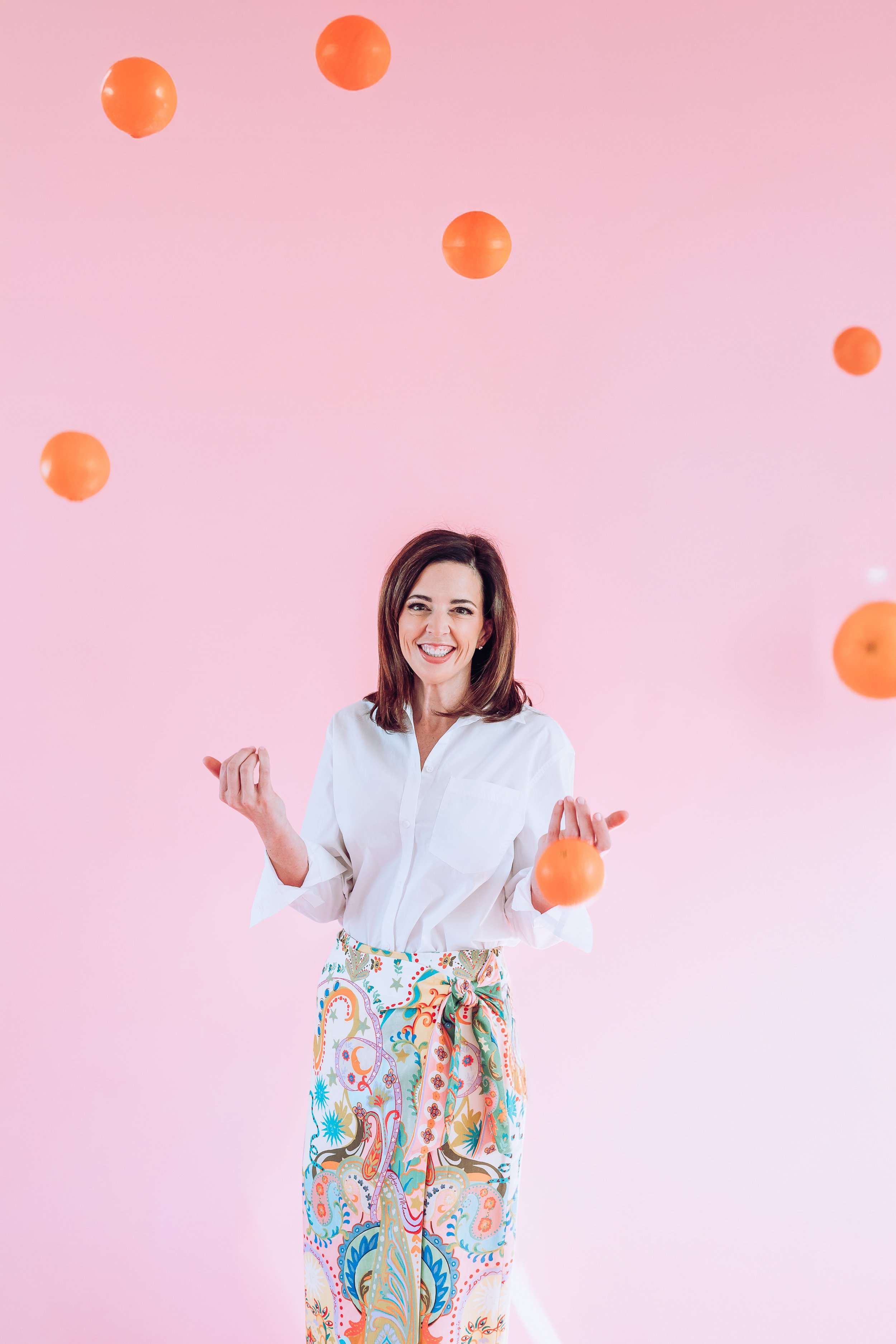 Warm, approachable female therapist standing against a soft pink background with floating oranges, conveying energy, optimism, and approachability in counseling.