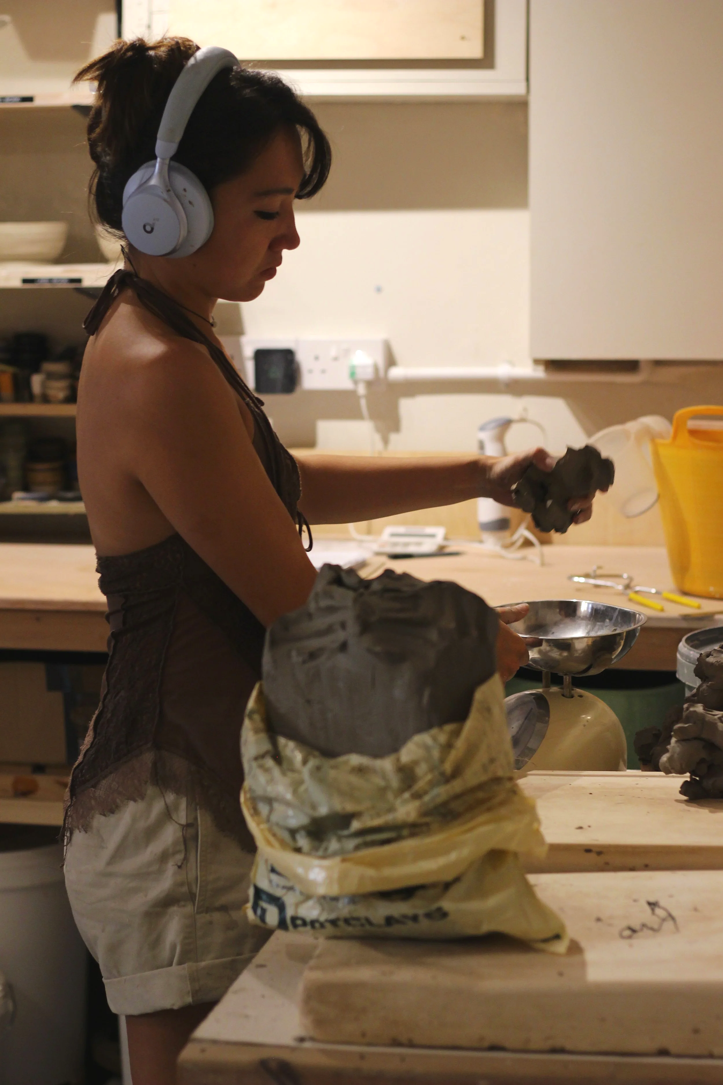 A woman wearing headphones, a sleeveless top, and shorts, working with clay at a workbench in a pottery studio.