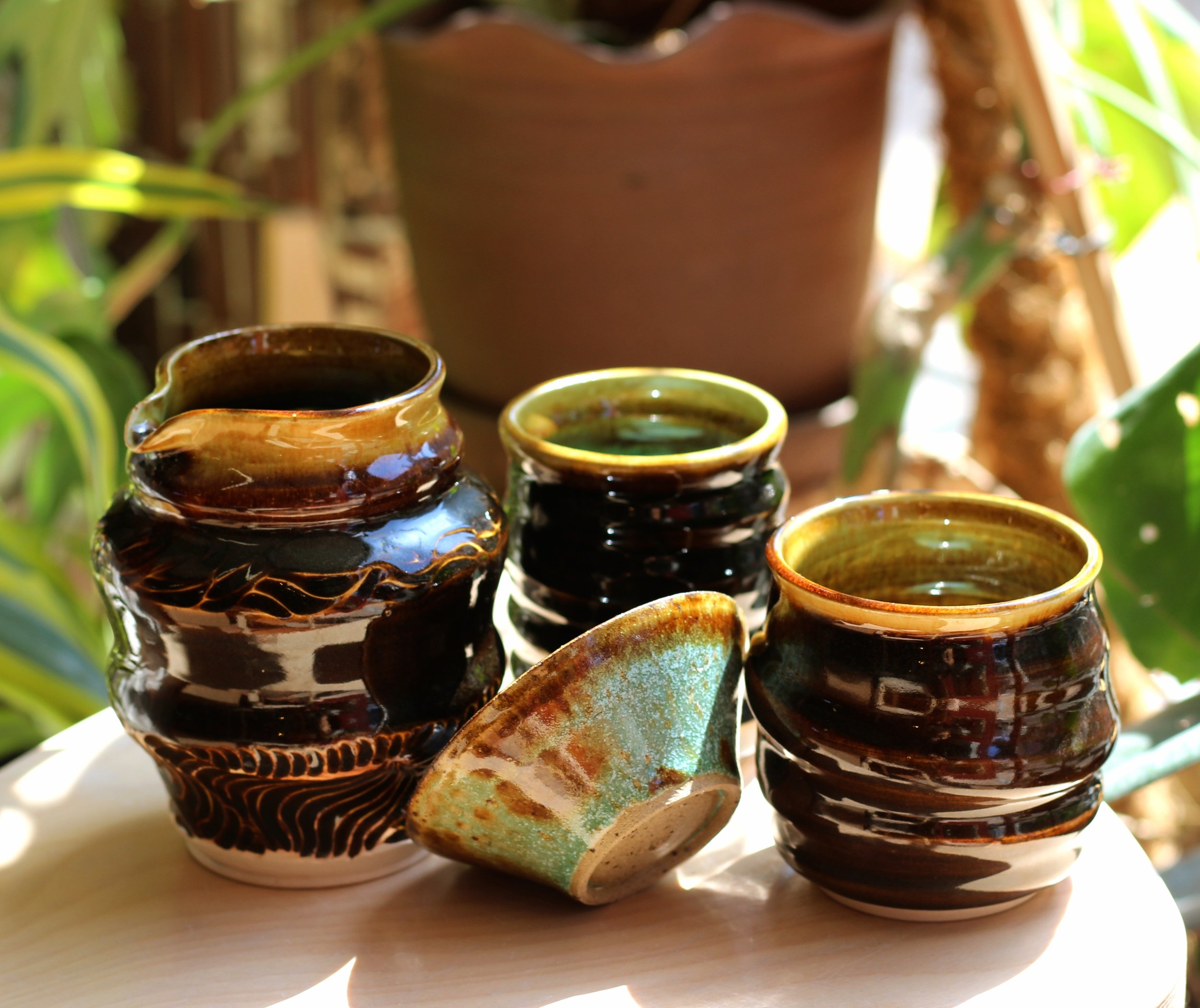 Three handcrafted ceramic cups and one small ceramic bowl on a light wooden surface, with green plants and a large potted plant in the background.