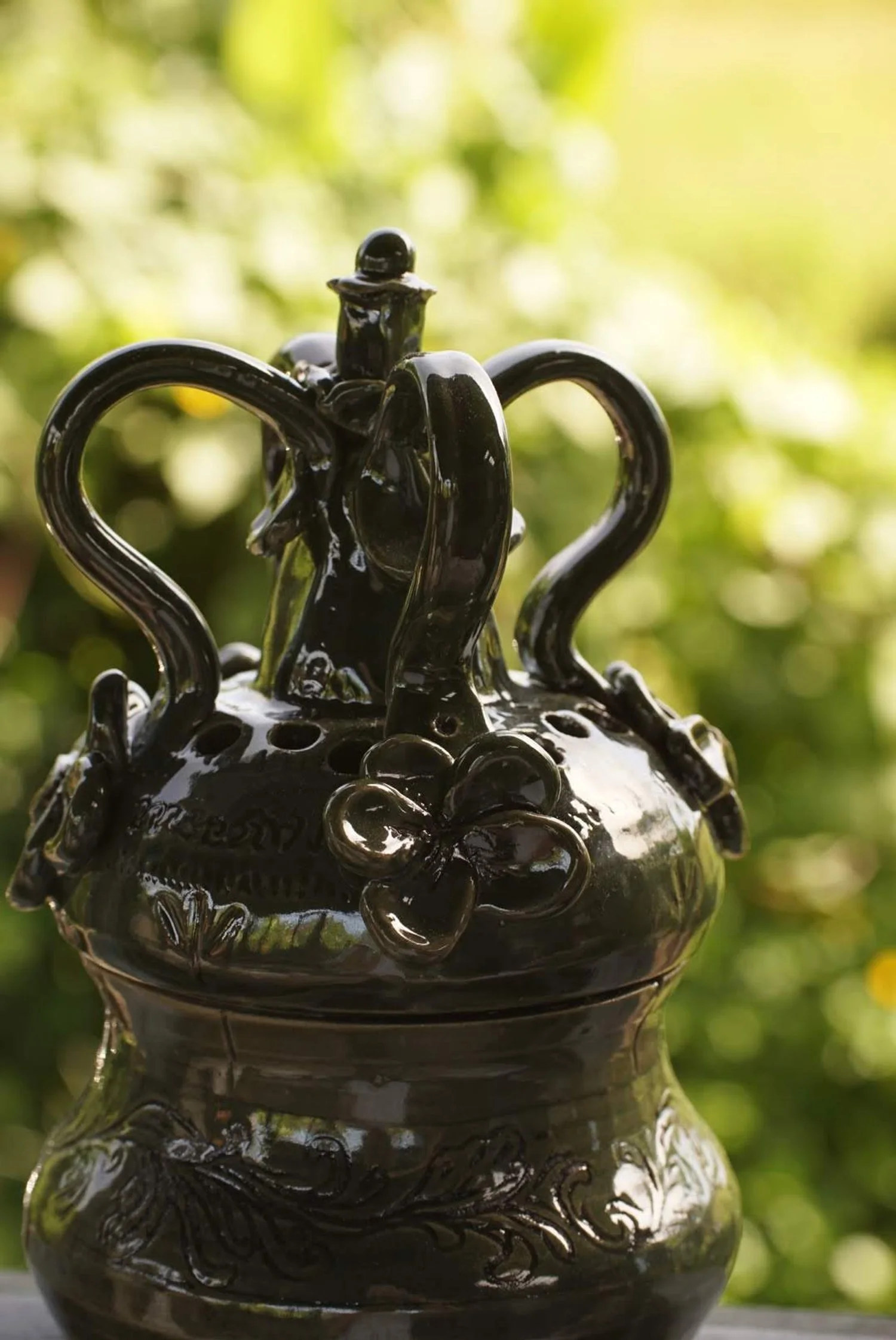 Black ceramic decorative vase with floral and swirl patterns, featuring a small flower embellishment and ornate handles, outdoors against a blurred green and yellow background.