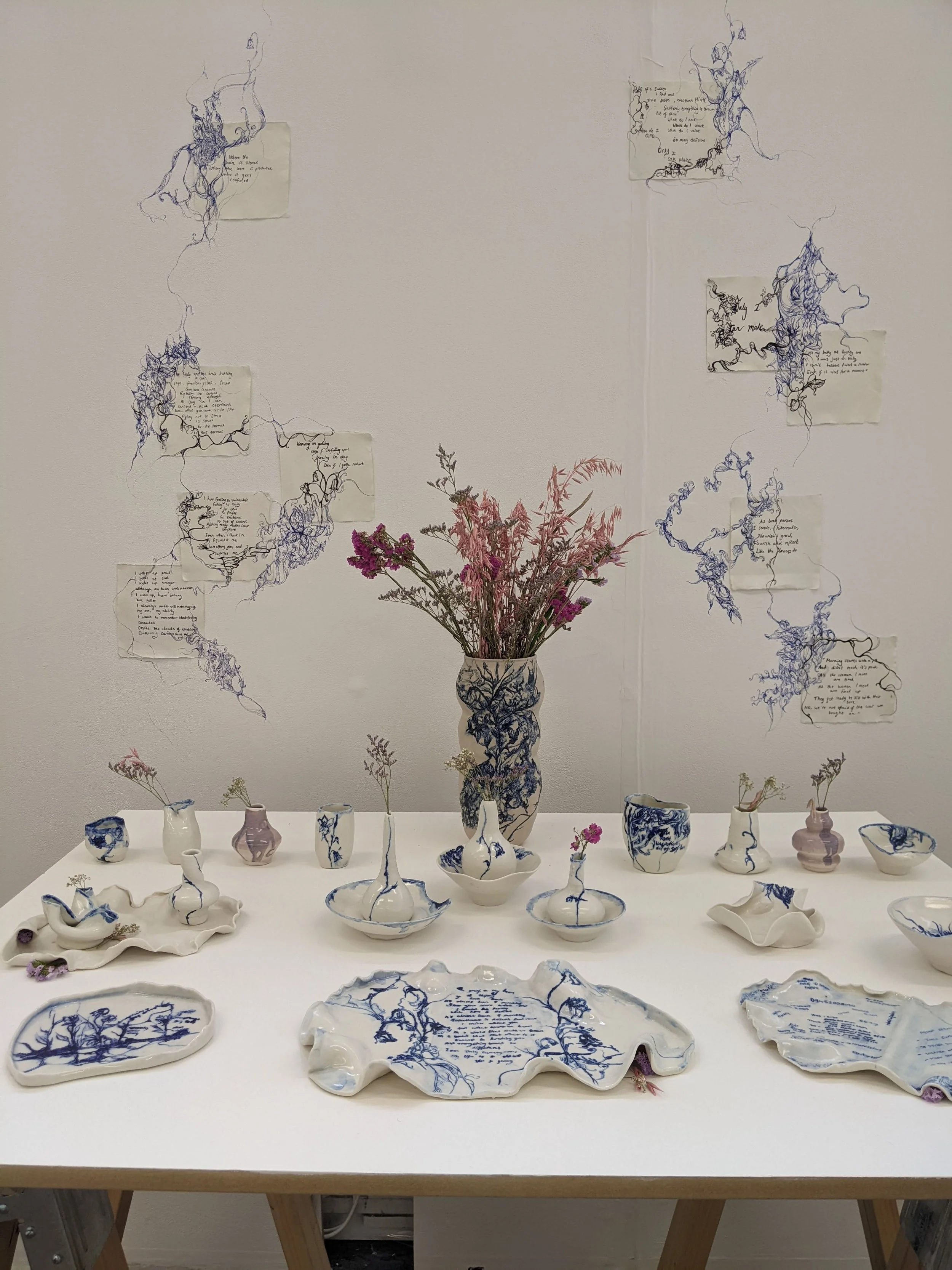An art installation featuring handmade ceramic vases and plates with blue painted tree and handwritten text designs. The ceramics are arranged on a white table, with dried flowers placed inside some of the vases. Flowers and handwritten poetry are also displayed on the wall behind. The wall and table are white, creating a minimalistic aesthetic.