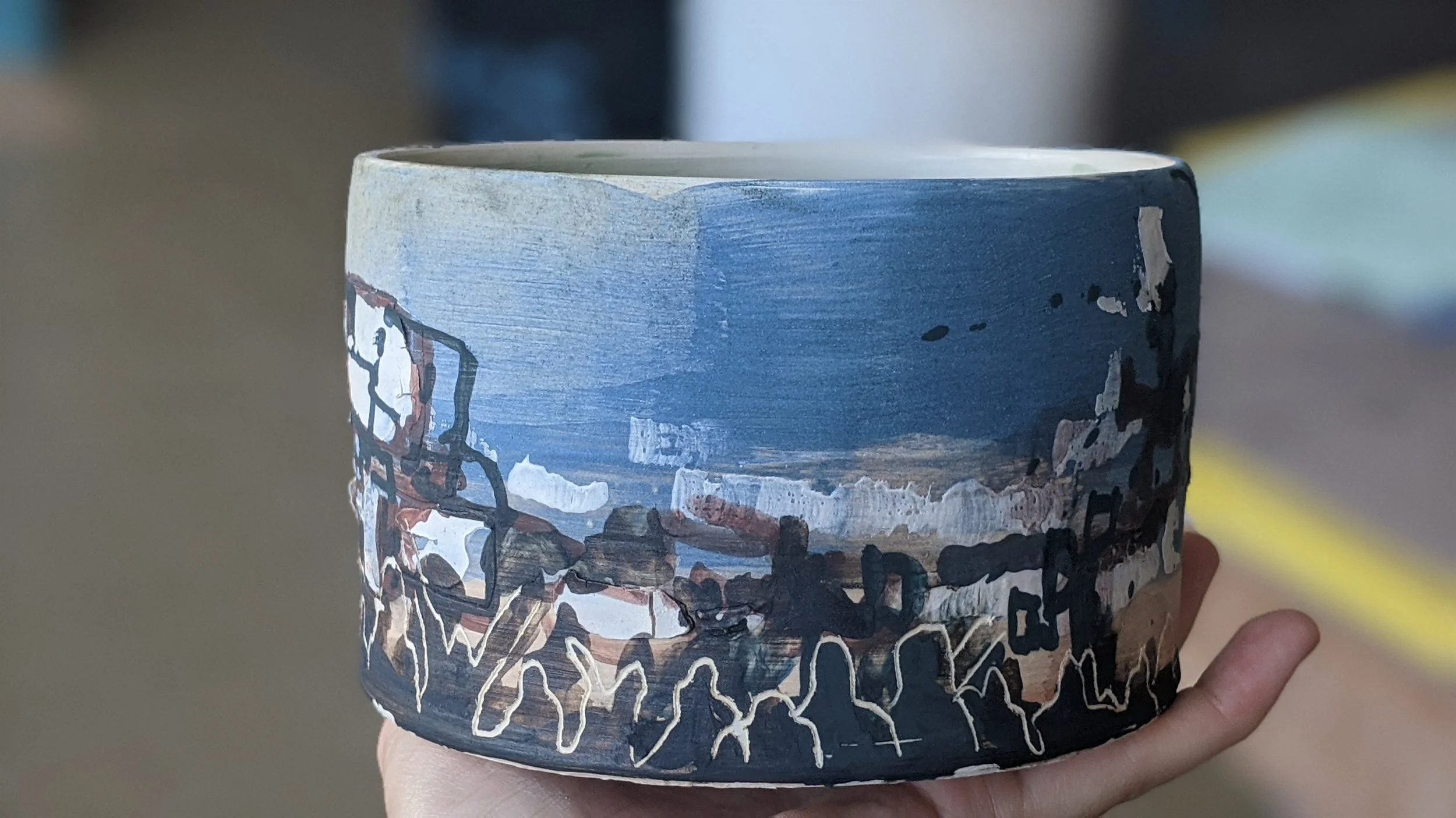 Hand holding a cylindrical ceramic mug painted with a landscape scene featuring blue sky, mountains, and black outlines of trees and buildings.