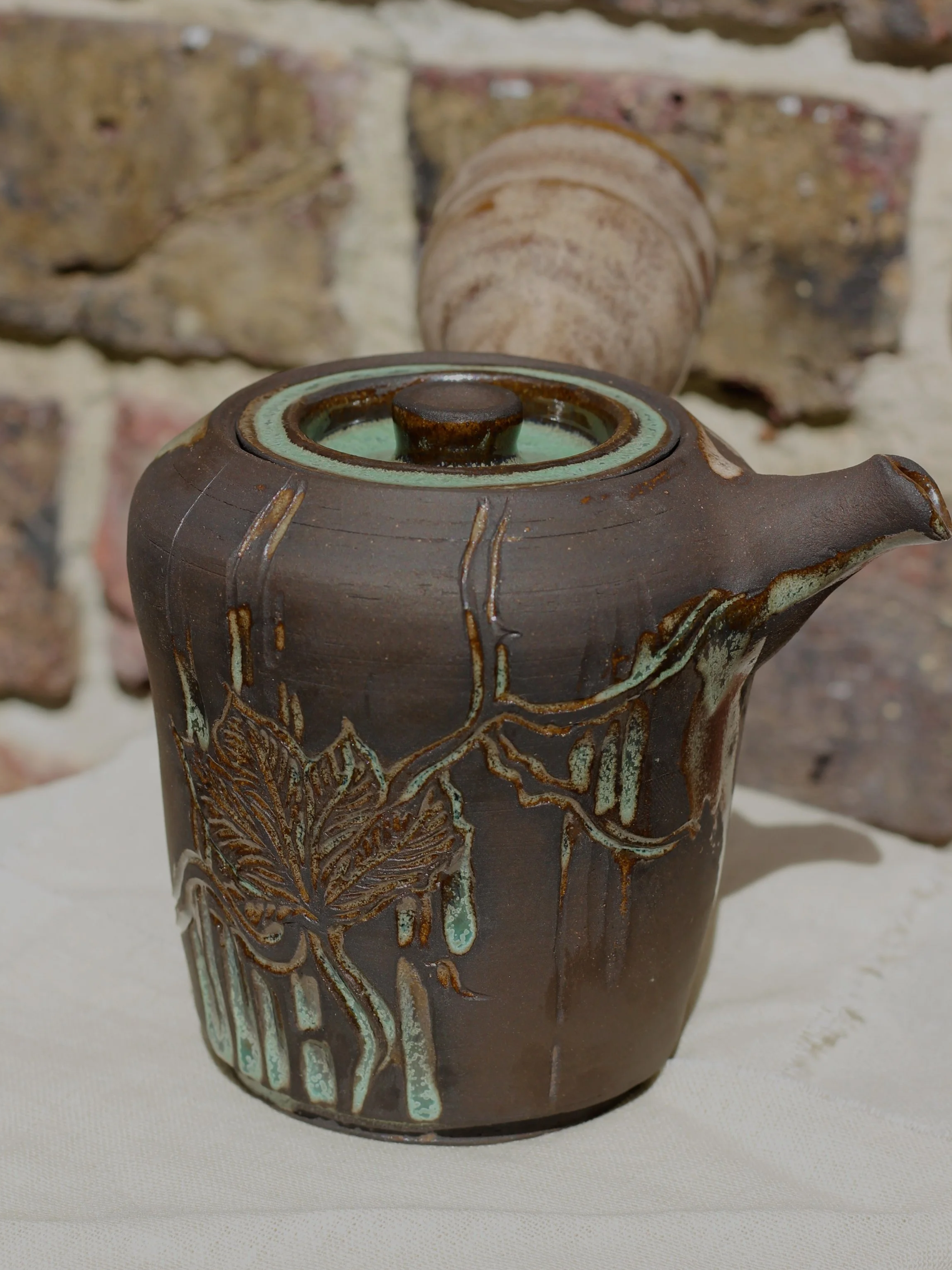 A ceramic teapot with artwork of a tree branch and leaves, featuring a textured, glazed finish, placed on a light-colored surface against a brick wall background.