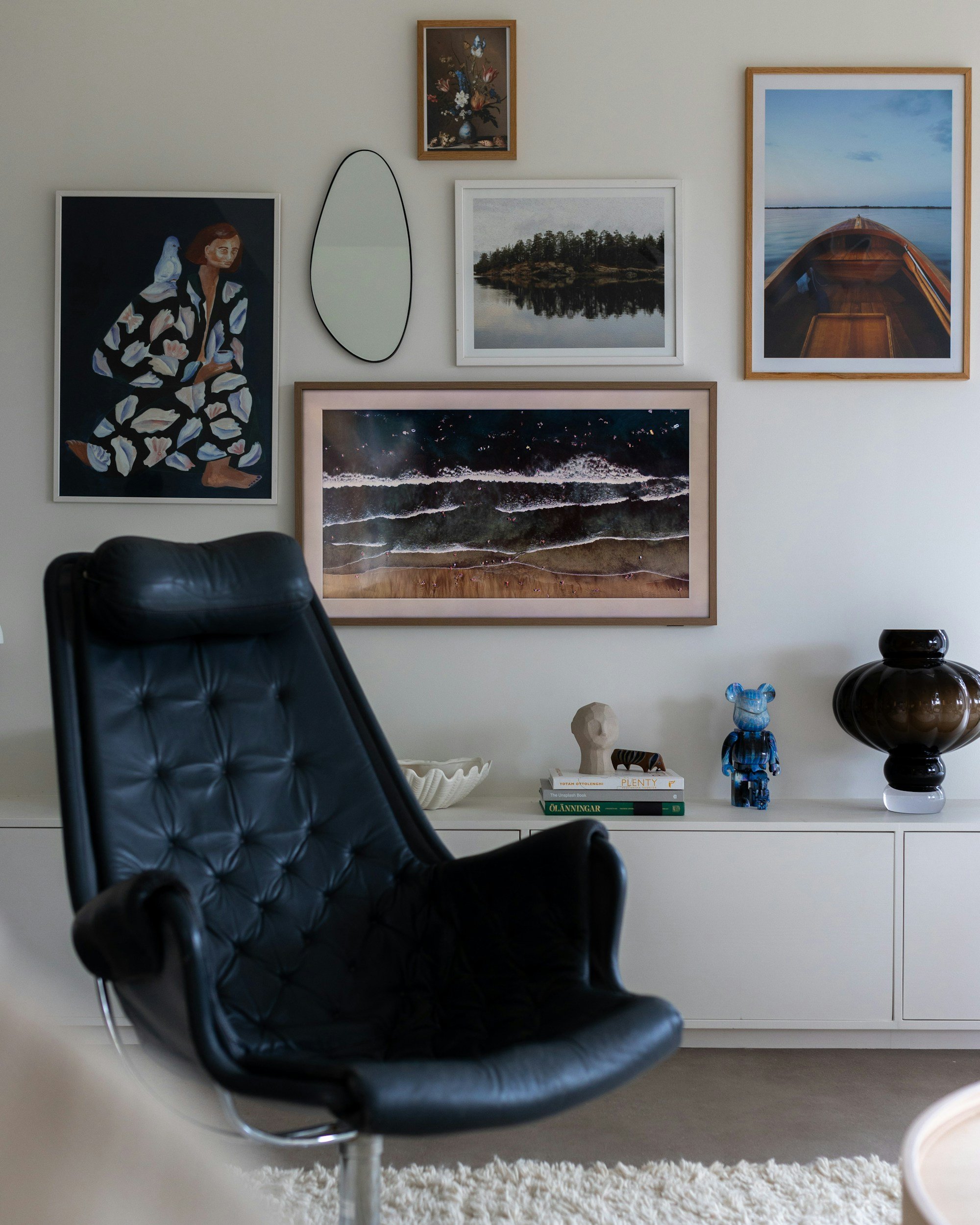 A black leather armchair in a living room with a white sideboard decorated with books, a sculpture, a blue glass figurine, and a dark vase. Above, a gallery wall features six framed artworks and a mirror.