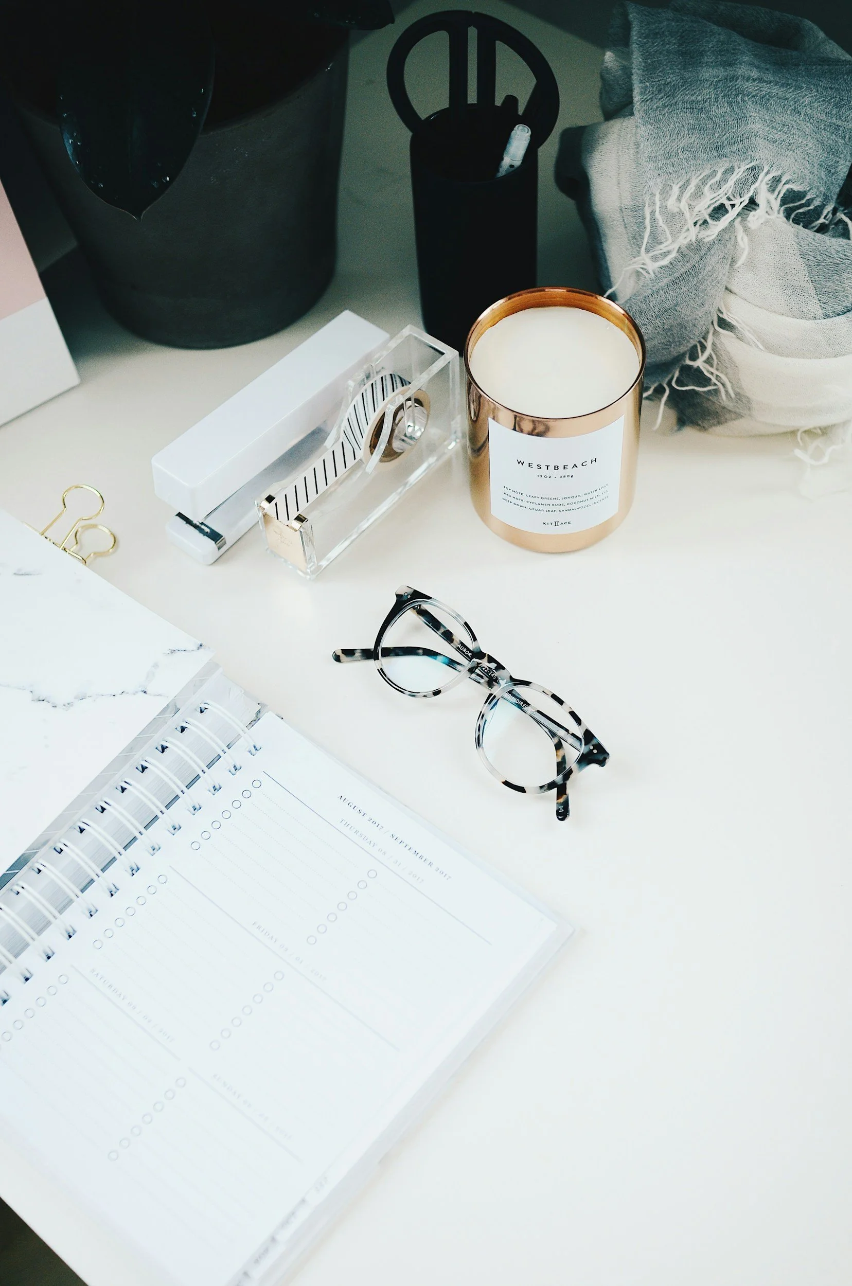 A workspace desk featuring a planner, a pair of tortoiseshell glasses, a candle, a notebook, a pen holder, a box cutter, and some paper clips.
