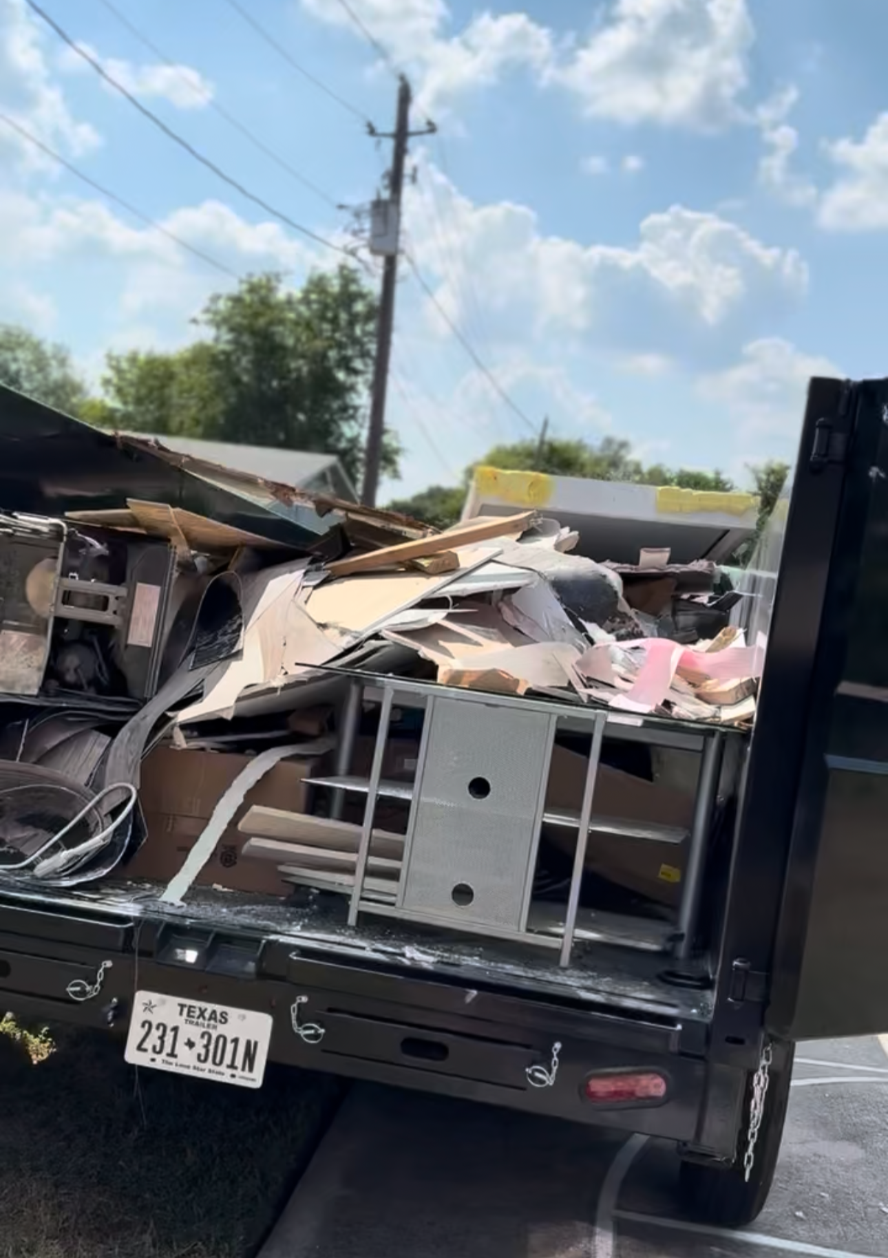 The back of a black flatbed truck filled with damaged office furniture, papers, and debris, parked on a road with a blue sky and some clouds in the background.