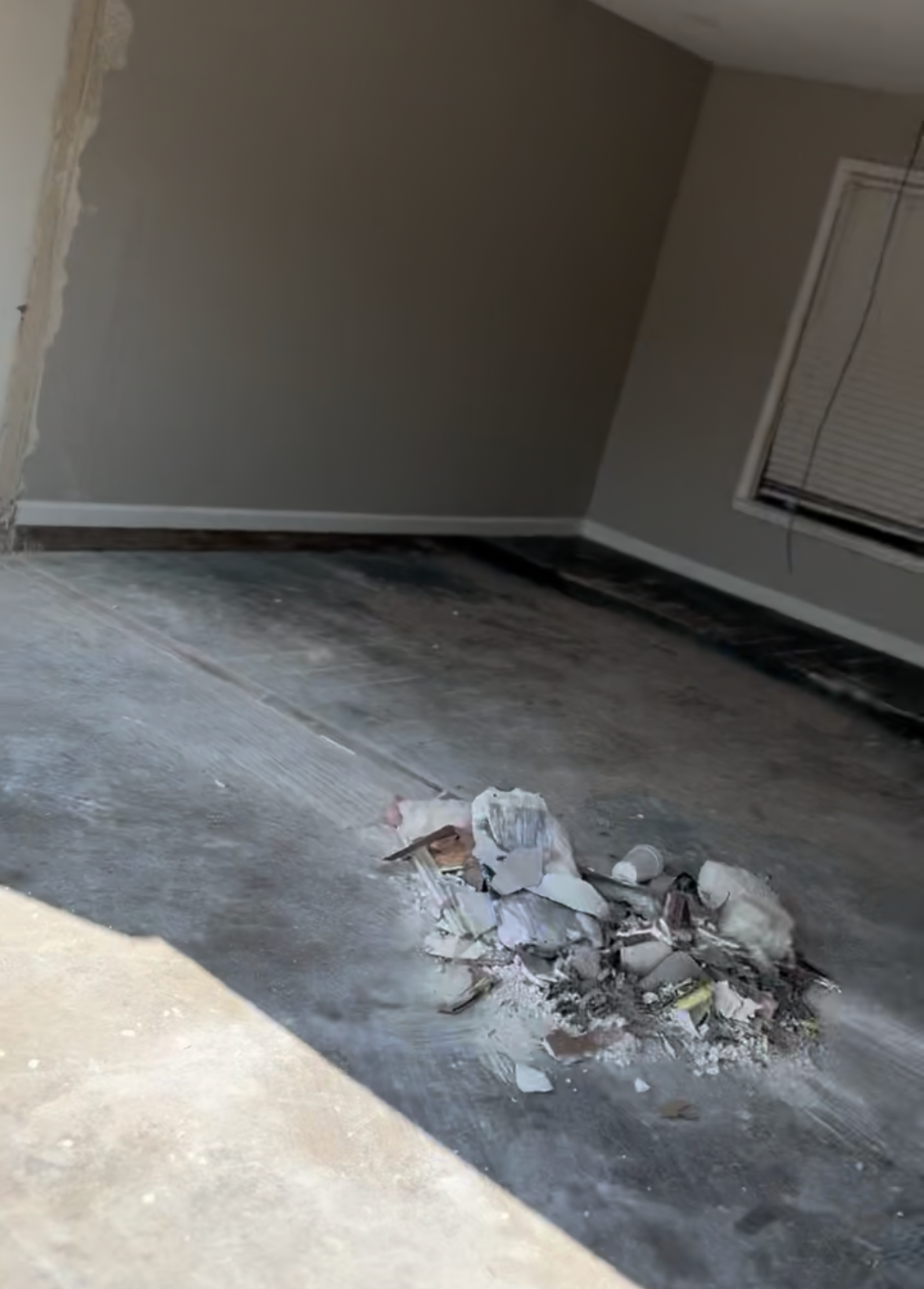 Debris and trash on a concrete floor inside a house near a wall and a window with closed blinds.