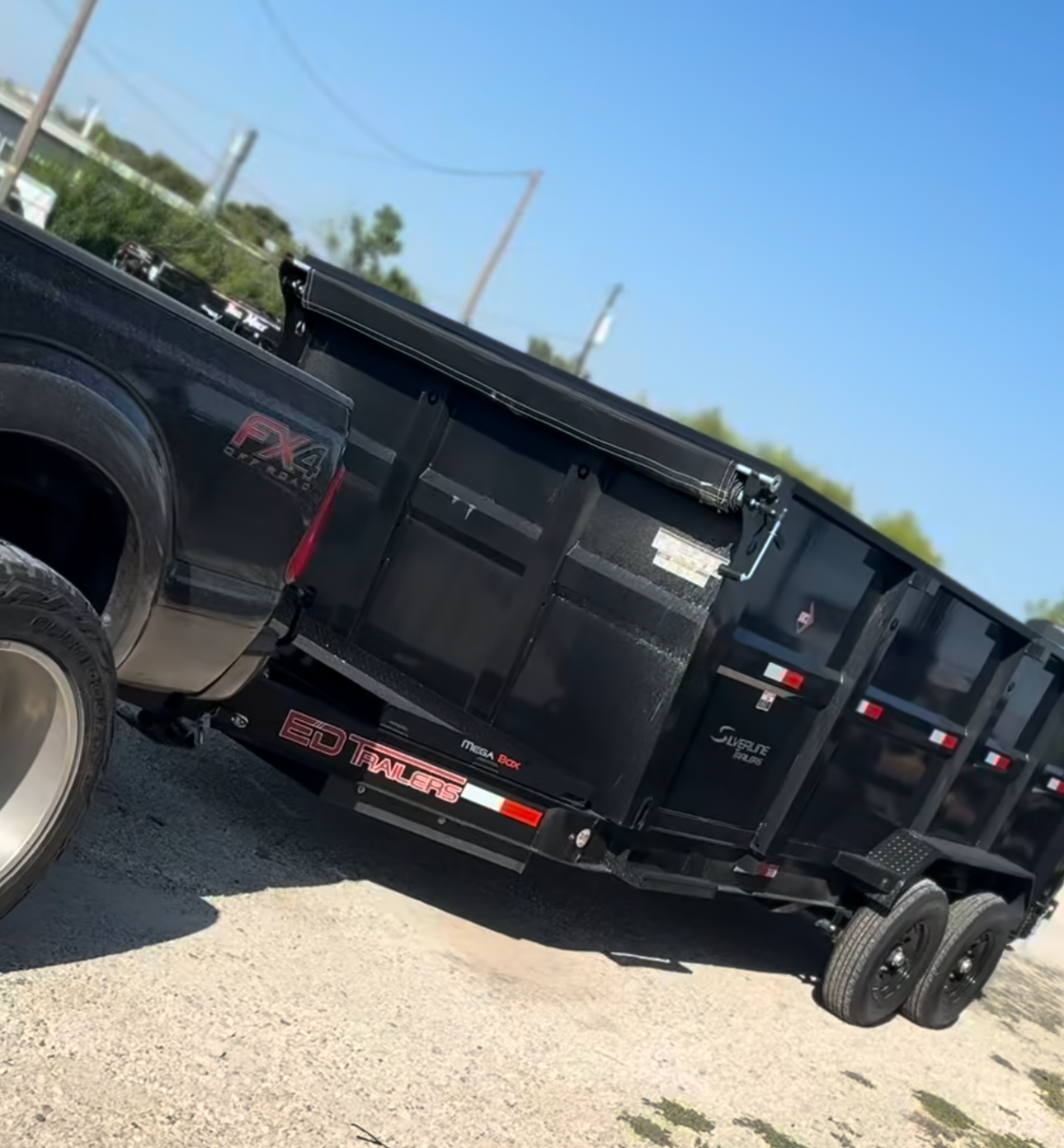 Black trailer with multiple wheels, labeled 'ED Trailers', parked on a gravel surface with blue sky and trees in the background.