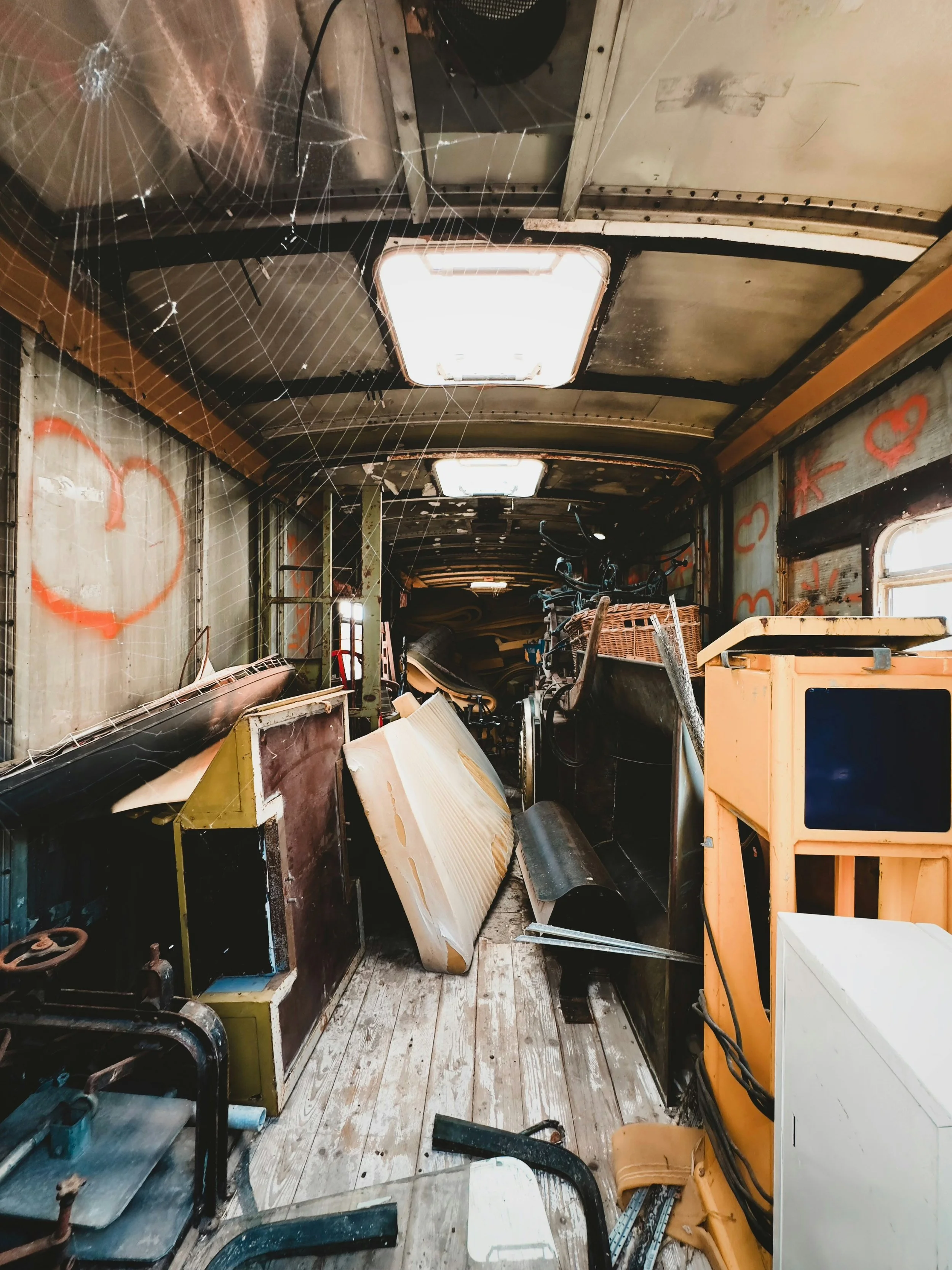 Interior of an abandoned, cluttered bus or train with cobwebs, graffiti on the walls, scattered debris, old furniture, bikes, and equipment.