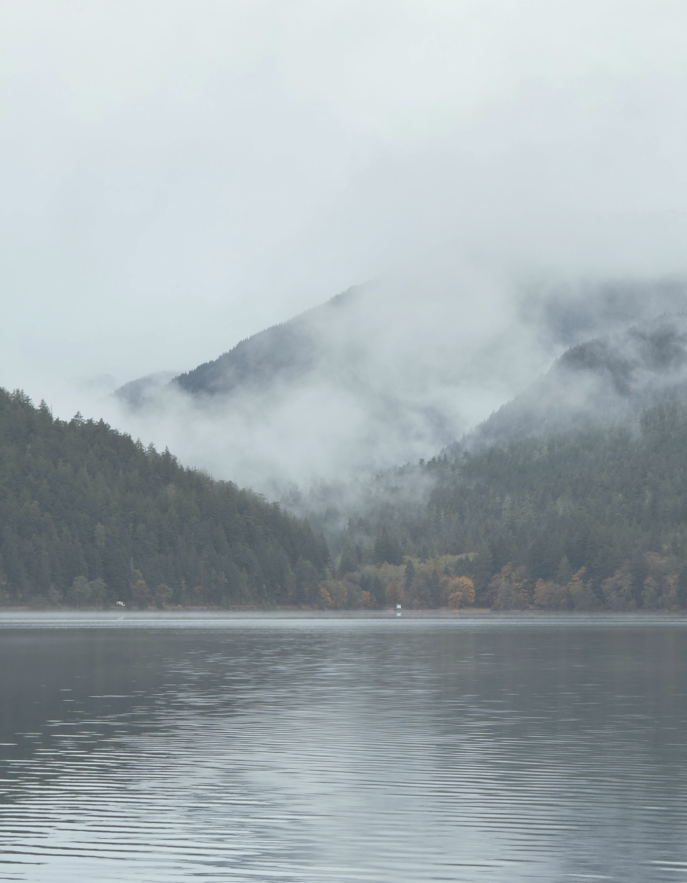 Mist over a calm lake with forested mountains in the background on an overcast day.
