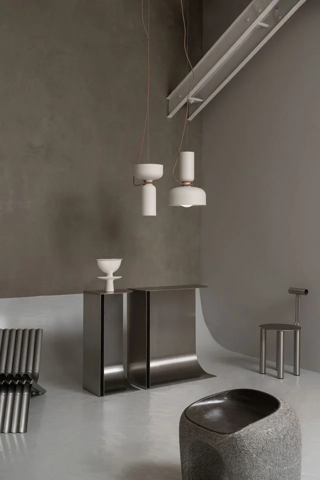 Décor minimaliste avec des suspensions blanches, une console en métal, une chaise métallique, une sculpture en pierre, et une surface en béton.