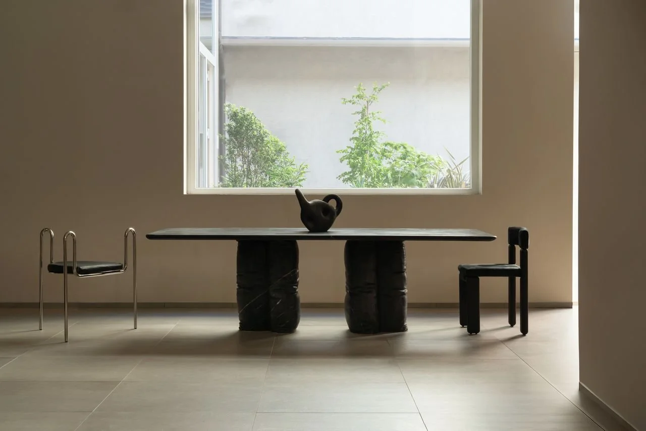 Une pièce minimaliste avec une grande fenêtre laissant entrer la lumière naturelle. Au centre, une table noire avec un objet en forme de cruche ou de vase. Deux chaises, l'une de style moderne avec une structure en métal et l'autre en bois noir, sont