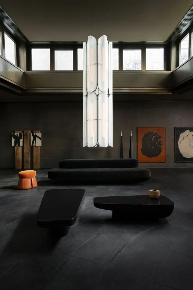 Intérieur moderne avec mobilier noir et une grande lampe suspendue, œuvres d'art abstraites au mur, et fenêtres en haut laissant entrer la lumière.