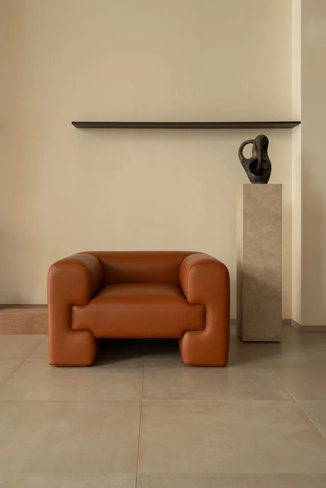 Un fauteuil en cuir marron clair, une sculpture noire sur un piédestal en pierre, une étagère simple et blanche contre un mur beige dans un espace minimaliste.
