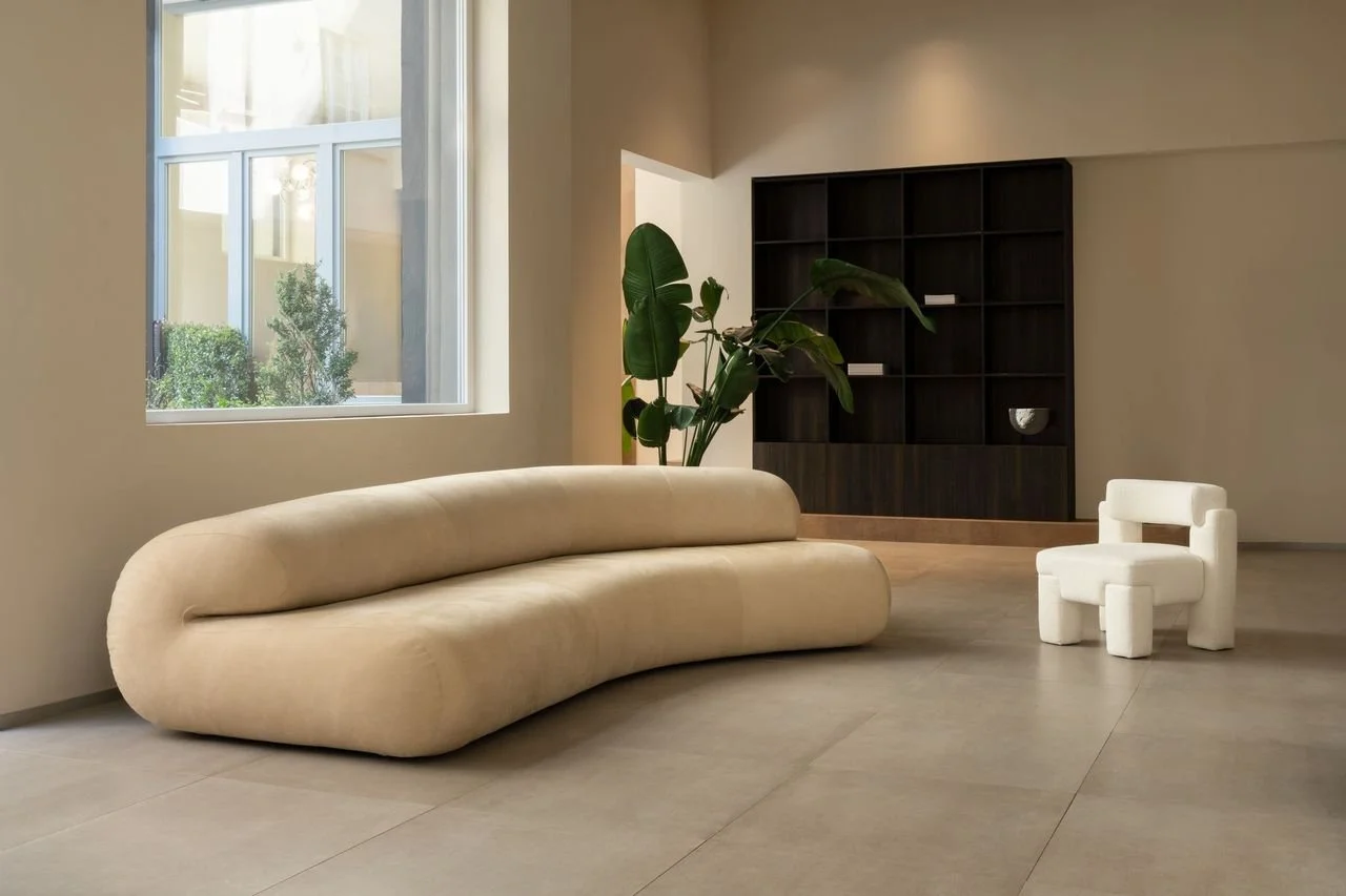 Un salon minimaliste avec un canapé beige, une petite chaise blanche, une grande plante verte, et une étagère noire en arrière-plan, à côté d'une fenêtre laissant entrer la lumière naturelle.