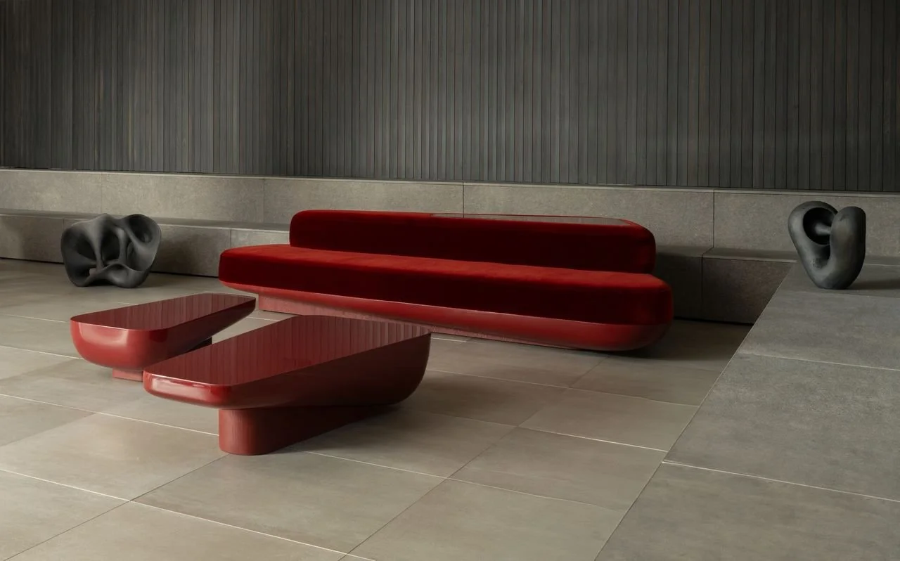 Un espace intérieur avec un canapé rouge, deux tables basses en bois, et deux sculptures abstraites noires, le tout sur un sol en carrelage gris devant un mur en panneaux gris foncé.